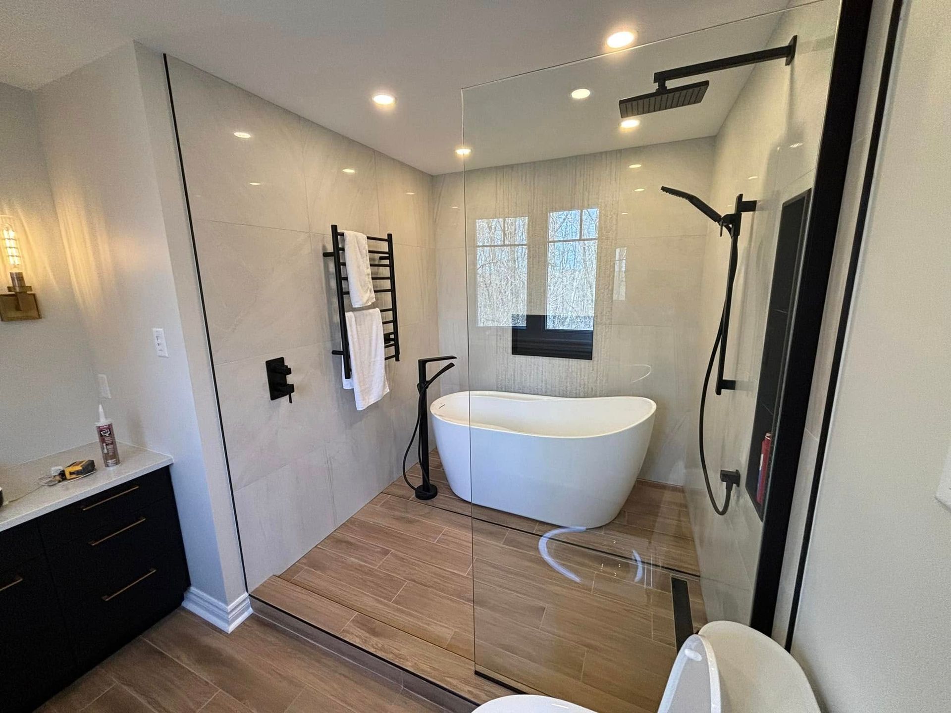 Une salle de bains moderne comprenant une baignoire blanche sur une estrade carrelée imitation bois, une douche vitrée et un meuble-lavabo noir.