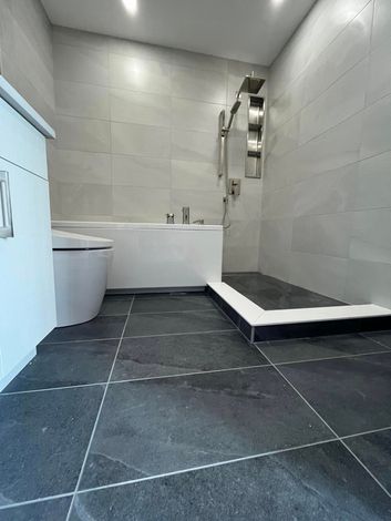 Une salle de bains moderne avec un sol carrelé en pierre gris foncé, une baignoire blanche et une douche à l'italienne carrelée de gris clair.