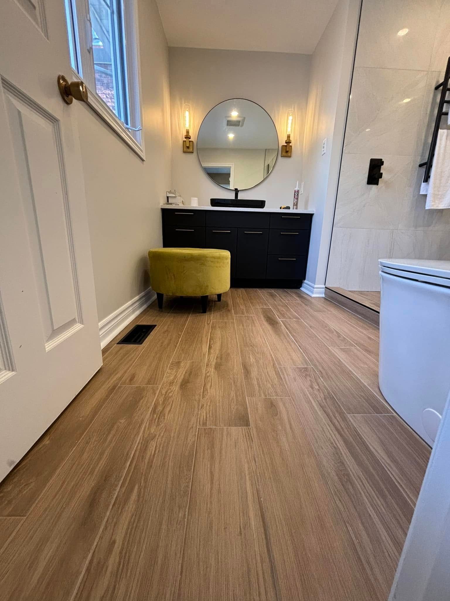 Une salle de bains moderne avec un sol imitation bois, un meuble-lavabo noir, un miroir rond et un pouf jaune.