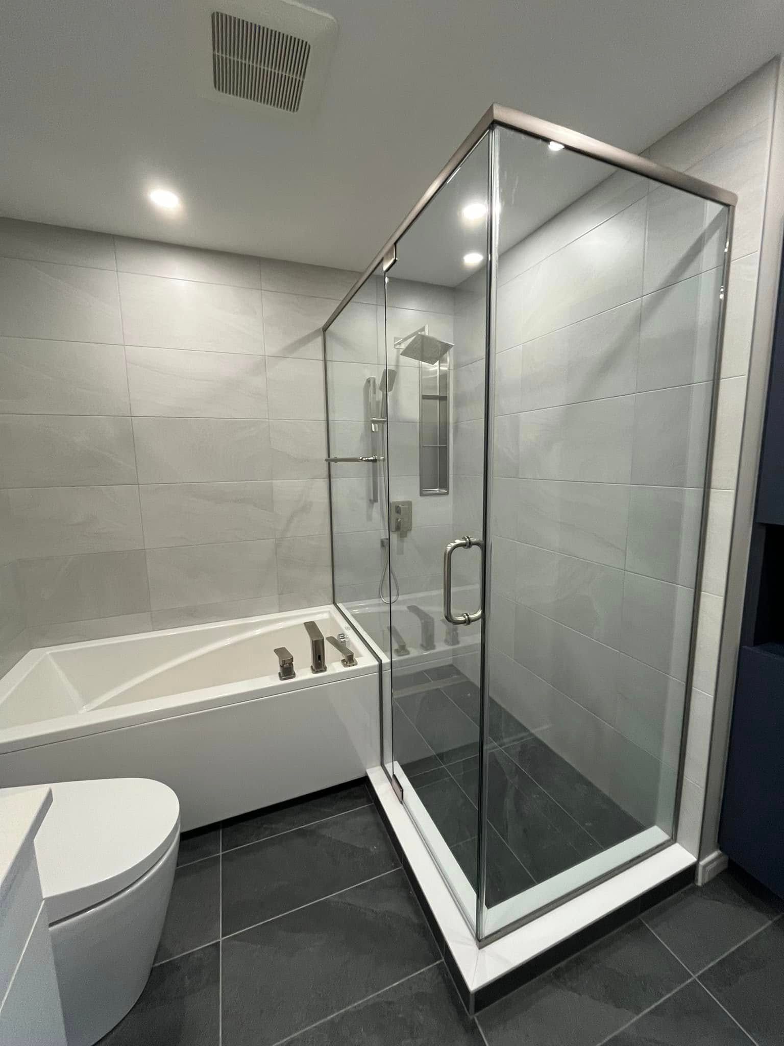 Une salle de bains moderne avec une baignoire blanche jouxtant une douche vitrée, des murs carrelés de gris et un sol carrelé de tons foncés.