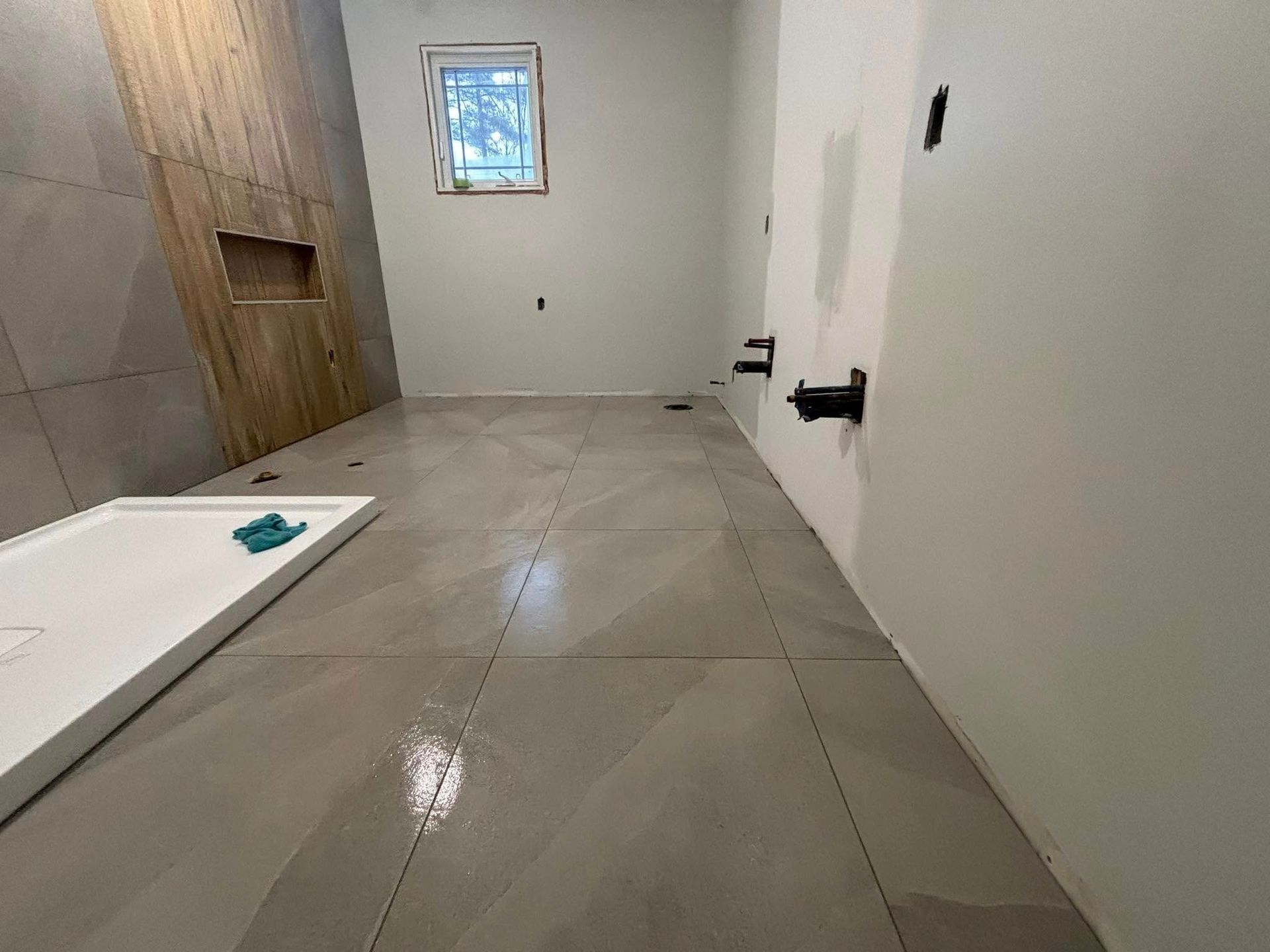 Une salle de bains en construction avec un sol carrelé gris clair, un receveur de douche blanc et des appareils sanitaires sur un mur blanc.