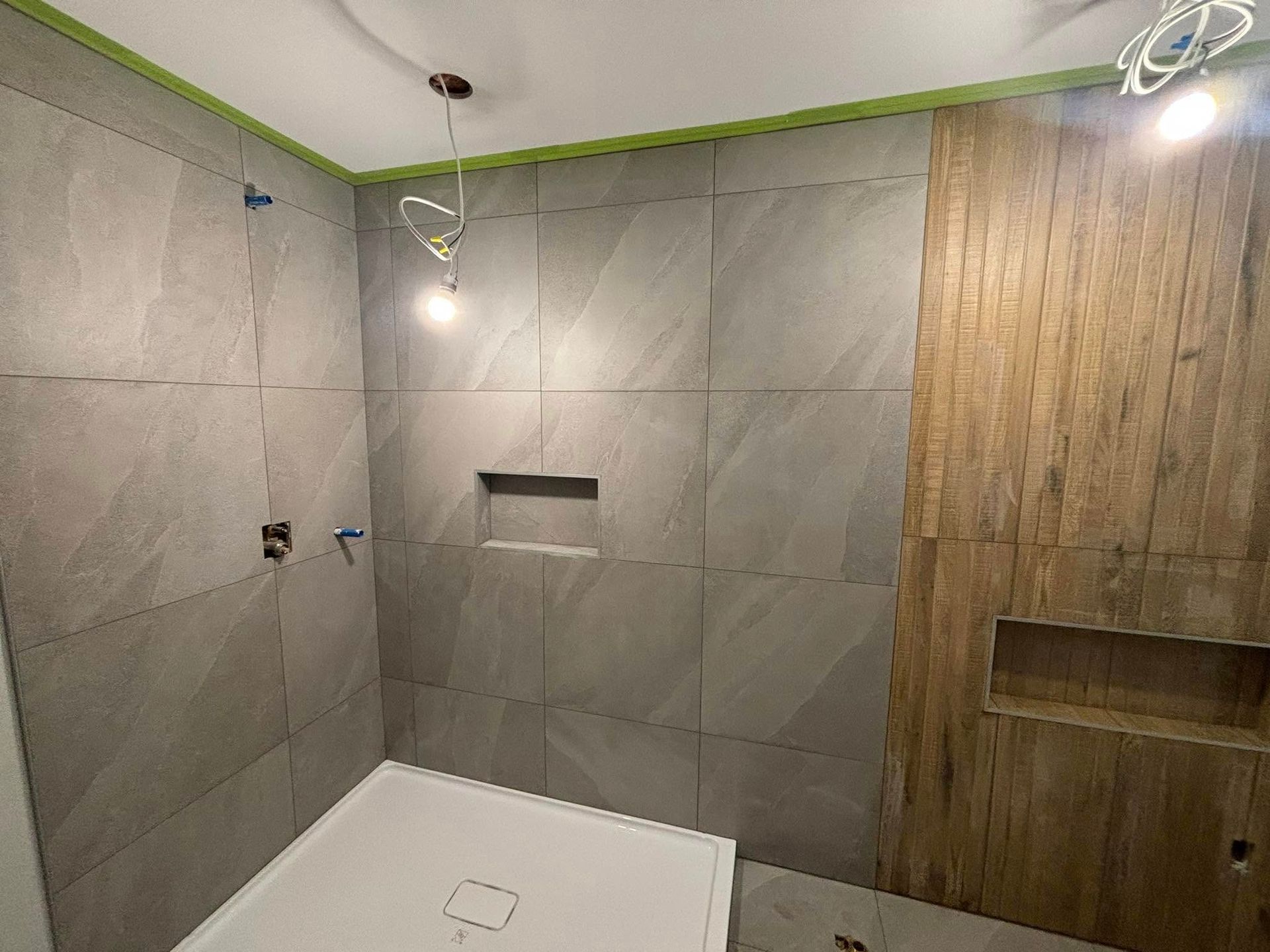 Une douche moderne en construction avec de grands carreaux gris, un mur d'accent carrelé imitation bois et deux étagères encastrées.
