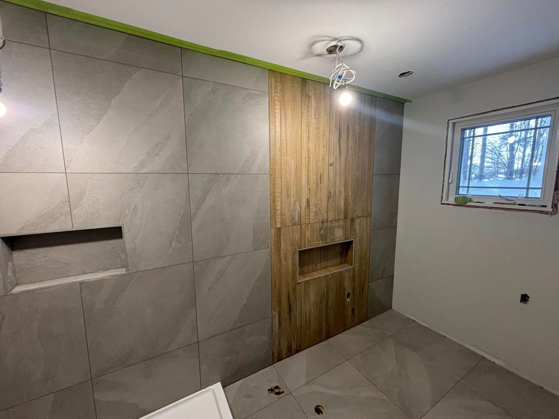 Une salle de bains en construction avec de grands carreaux muraux gris imitation pierre, une frise murale verticale en carrelage imitation bois et une fenêtre.