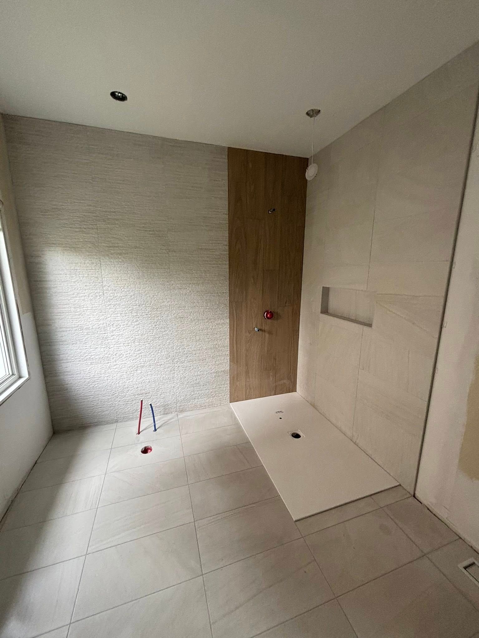 Une salle de bains inachevée avec de grands carreaux de sol gris, des carreaux muraux texturés gris clair et un accent de carreaux verticaux imitation bois.