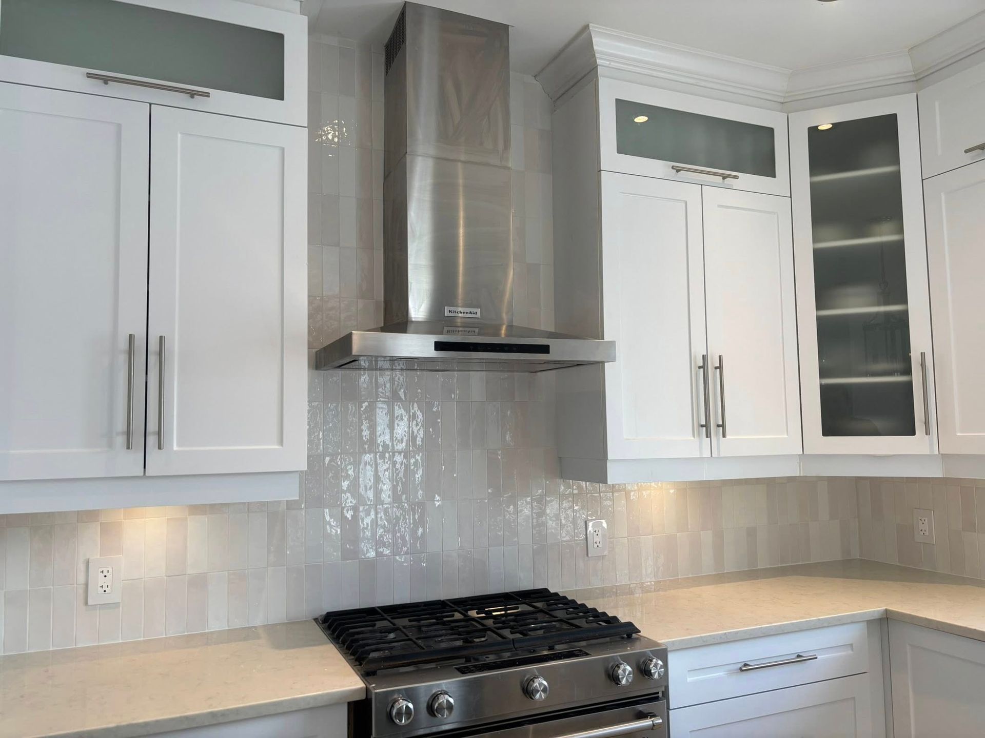Une hotte en acier inoxydable est installée au-dessus d'une plaque de cuisson, entre des armoires de cuisine blanches dont les portes supérieures sont vitrées.