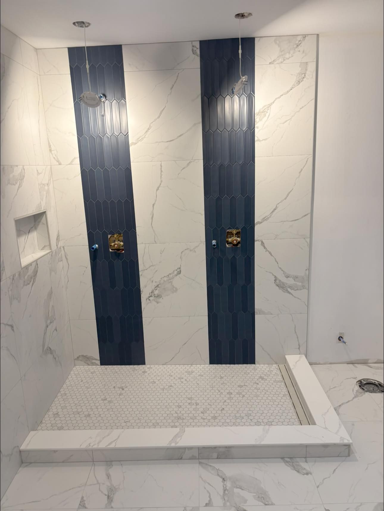 Une douche à l'italienne moderne avec des carreaux de marbre blanc, deux panneaux verticaux bleu marine, une robinetterie dorée et un sol en galets.