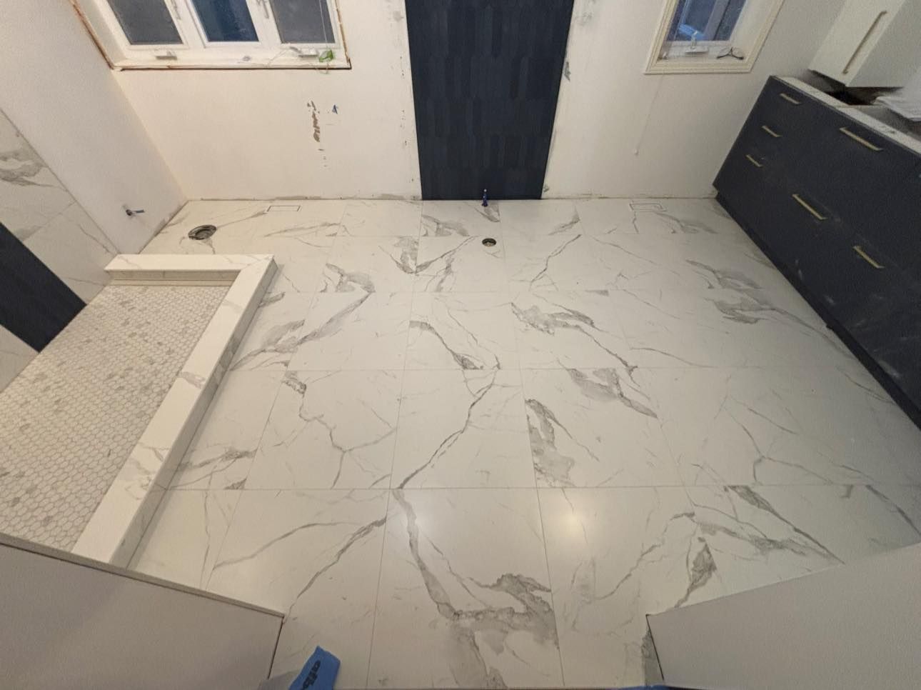 Vue en plongée d'un sol de salle de bains avec des carreaux à motifs de marbre blanc, un receveur de douche carrelé et des meubles sombres.