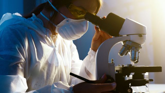 Person in protective gear looking through a microscope in a laboratory.