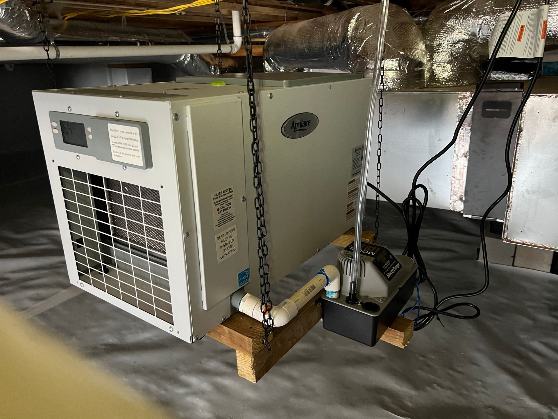 An air conditioner unit suspended in a crawlspace. A condensate pump is to its right.
