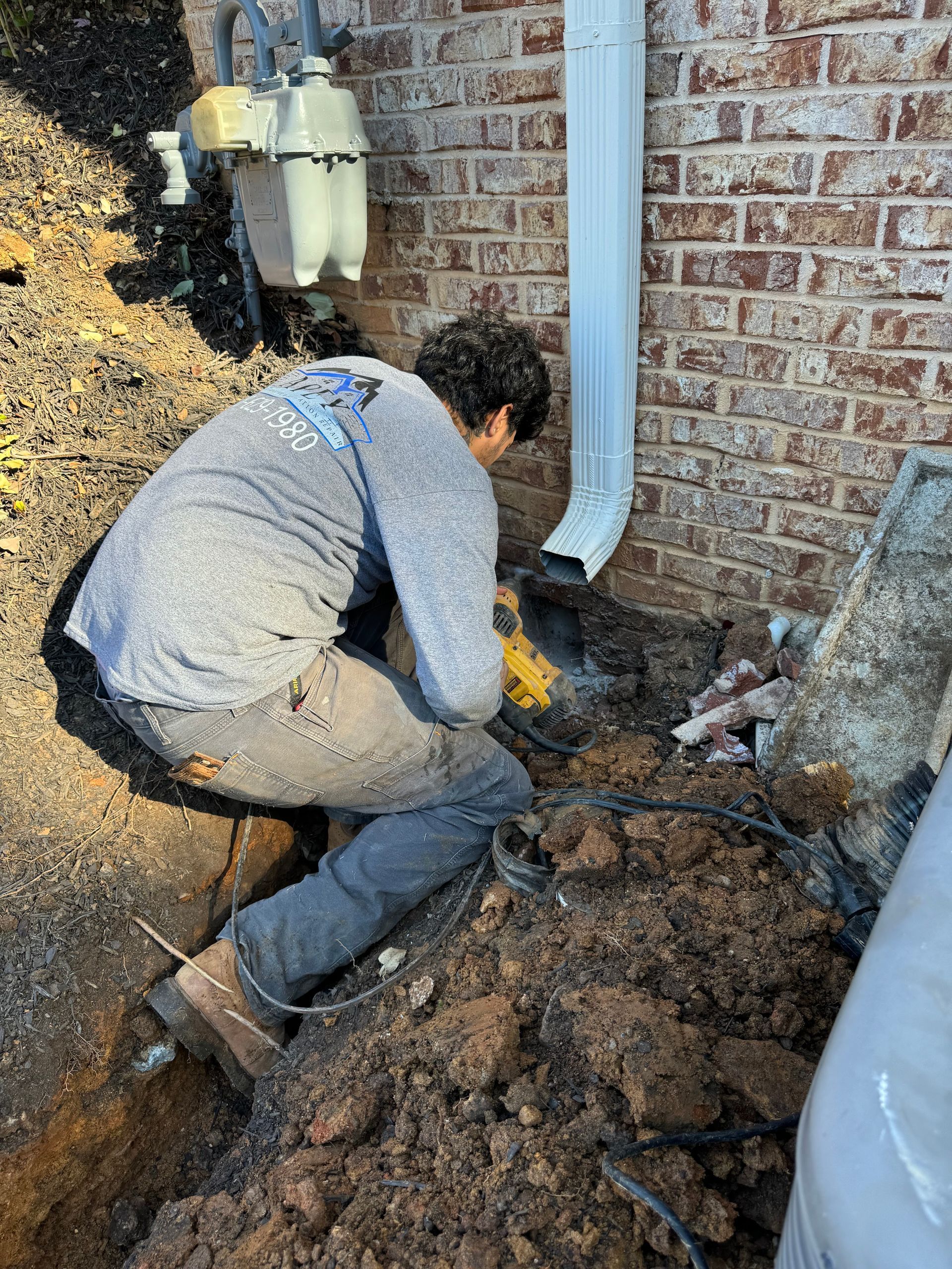 Person crouched, working near a brick wall and gas meter. Digging in dirt near a downspout with a power tool.