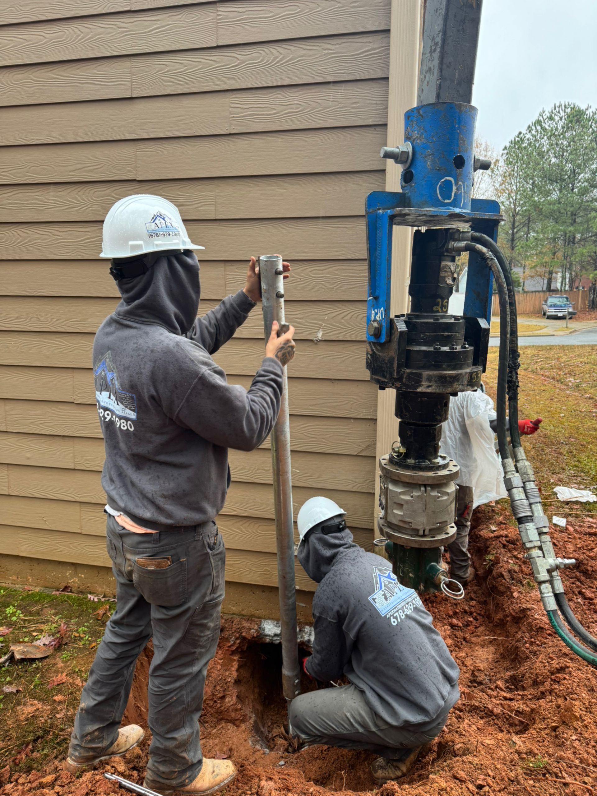 Two workers installing foundation piers next to a building. One operates a hydraulic machine, another uses a tool.