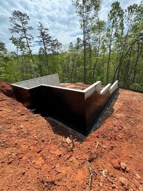 Concrete foundation walls on a red dirt hillside, surrounded by trees under a cloudy sky.