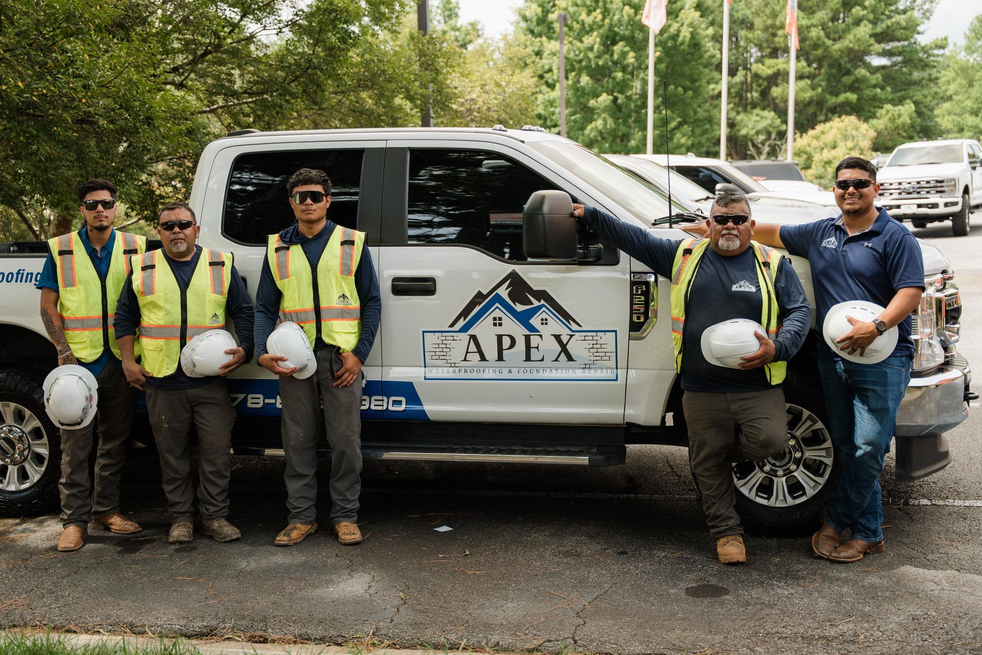 Five workers in safety vests and hard hats pose beside a white APEX utility truck outdoors.