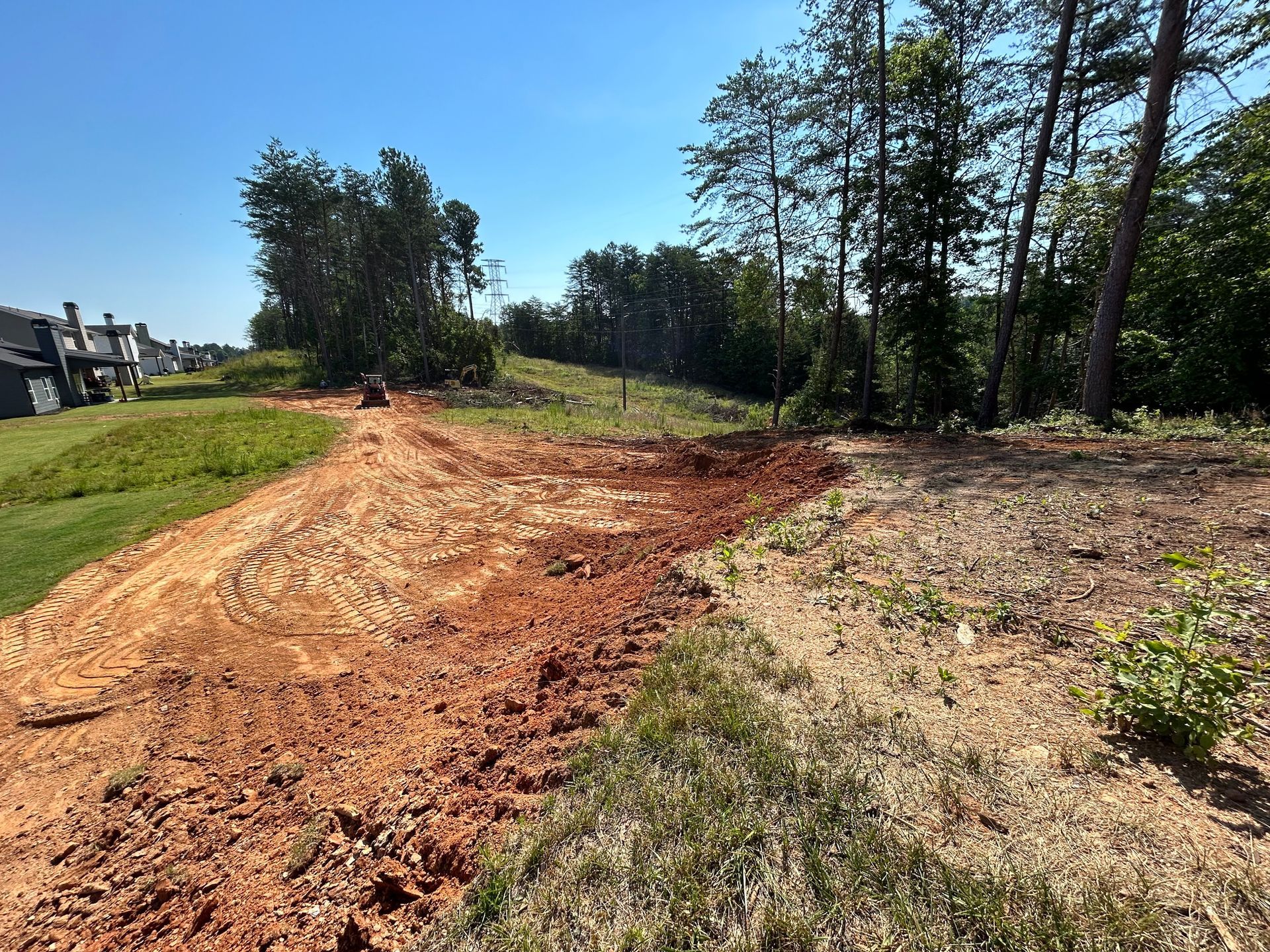 A construction site with exposed red soil, grass, trees, and houses under a blue sky.