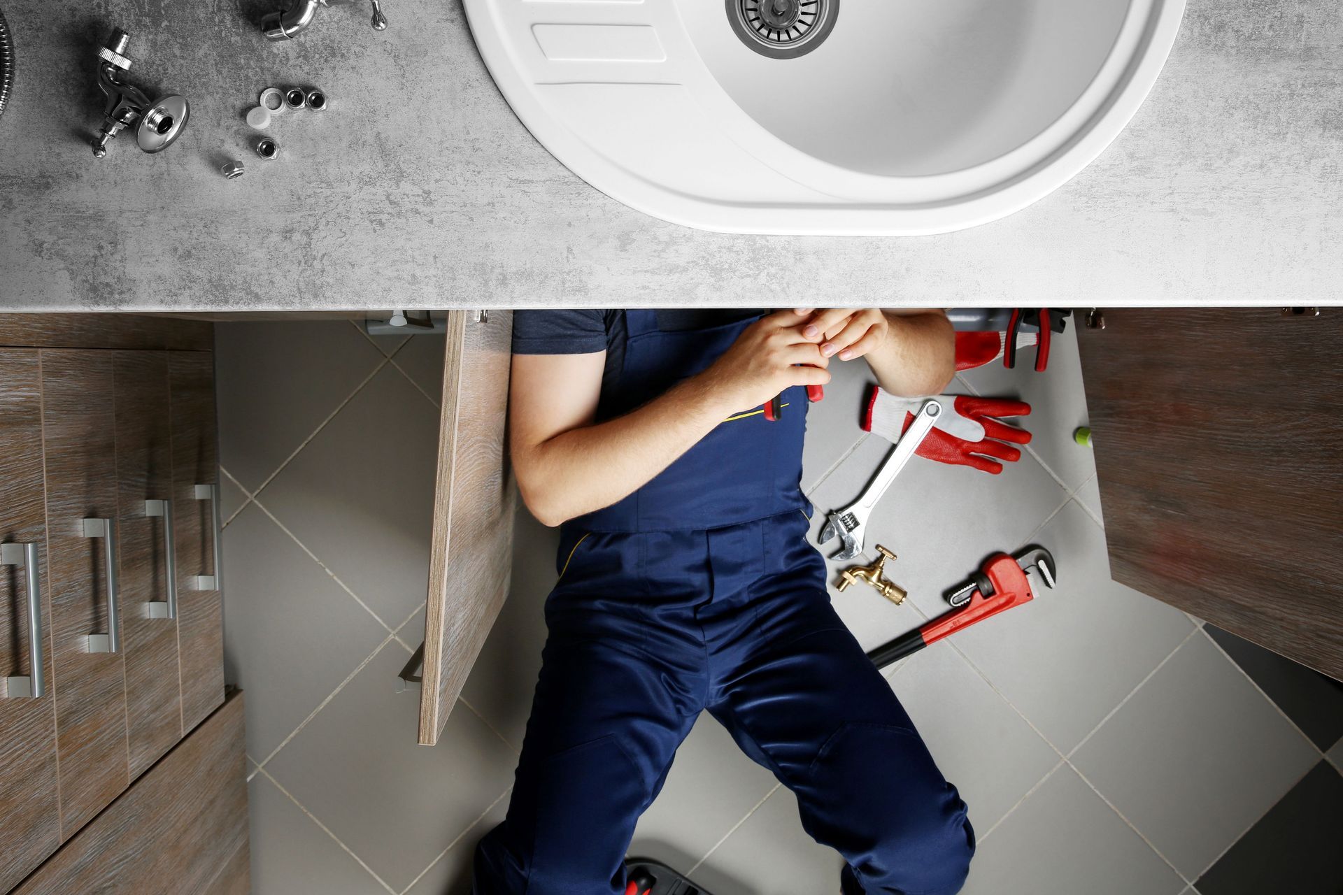 Person in blue overalls fixing sink pipes under bathroom cabinet with tools scattered.