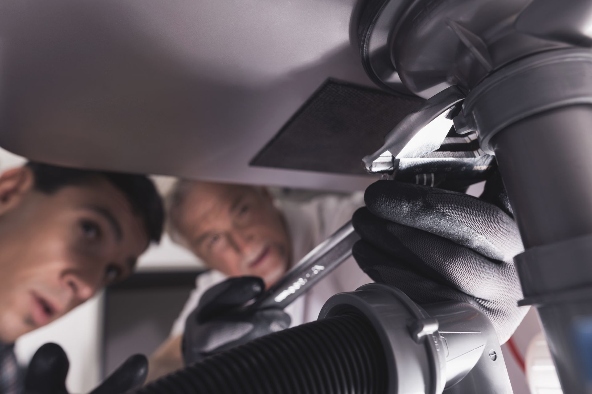 a close-up of a person wearing black gloves using a wrench to tighten plumbing pipes under a sink.