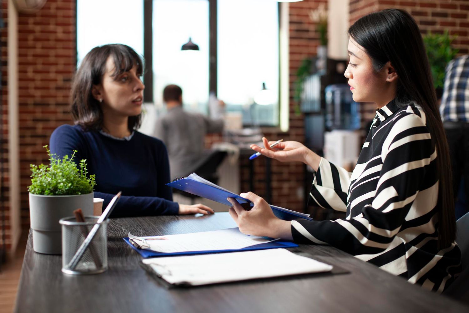Two people discuss documents at a table in a bright office or cafe.