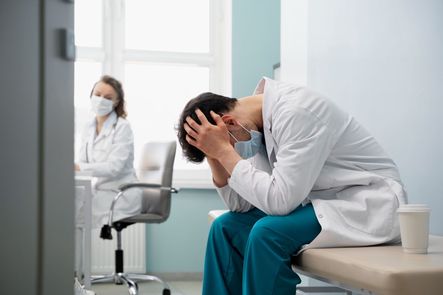 Distressed patient in white coat sits hunched with head in hands in a clinic, while a masked clinician sits nearby