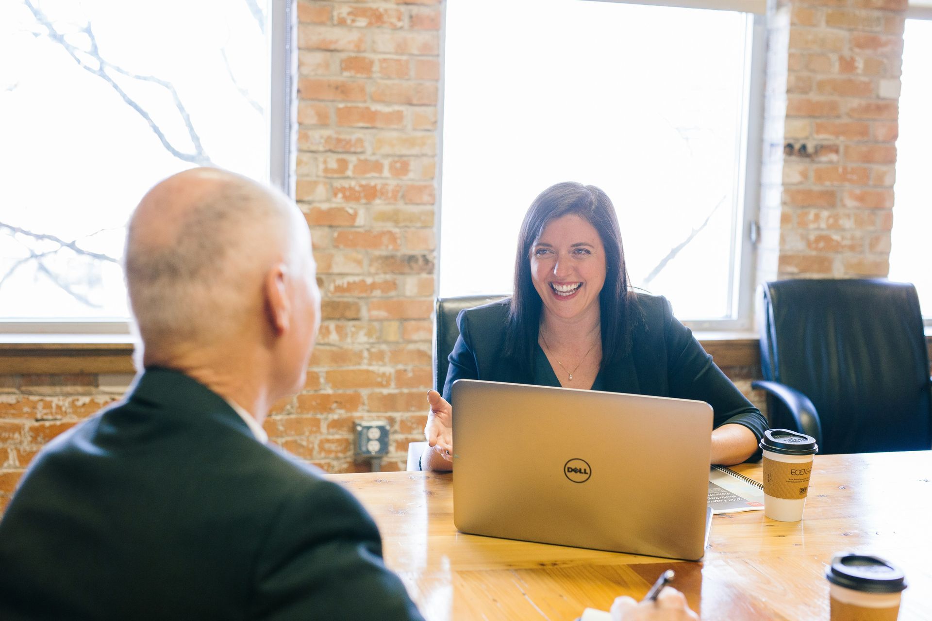 Two people in a meeting, one smiling behind a laptop at a conference table in a bright office.