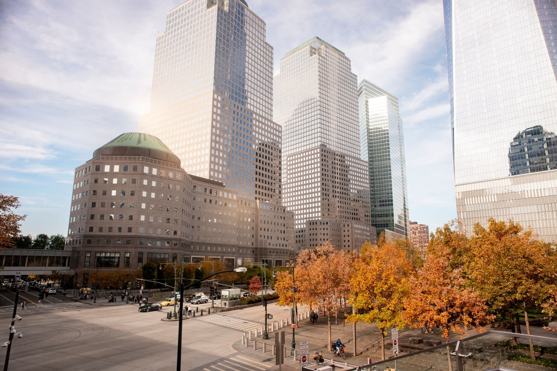 Downtown city skyline with tall glass buildings, autumn trees, and a busy plaza below