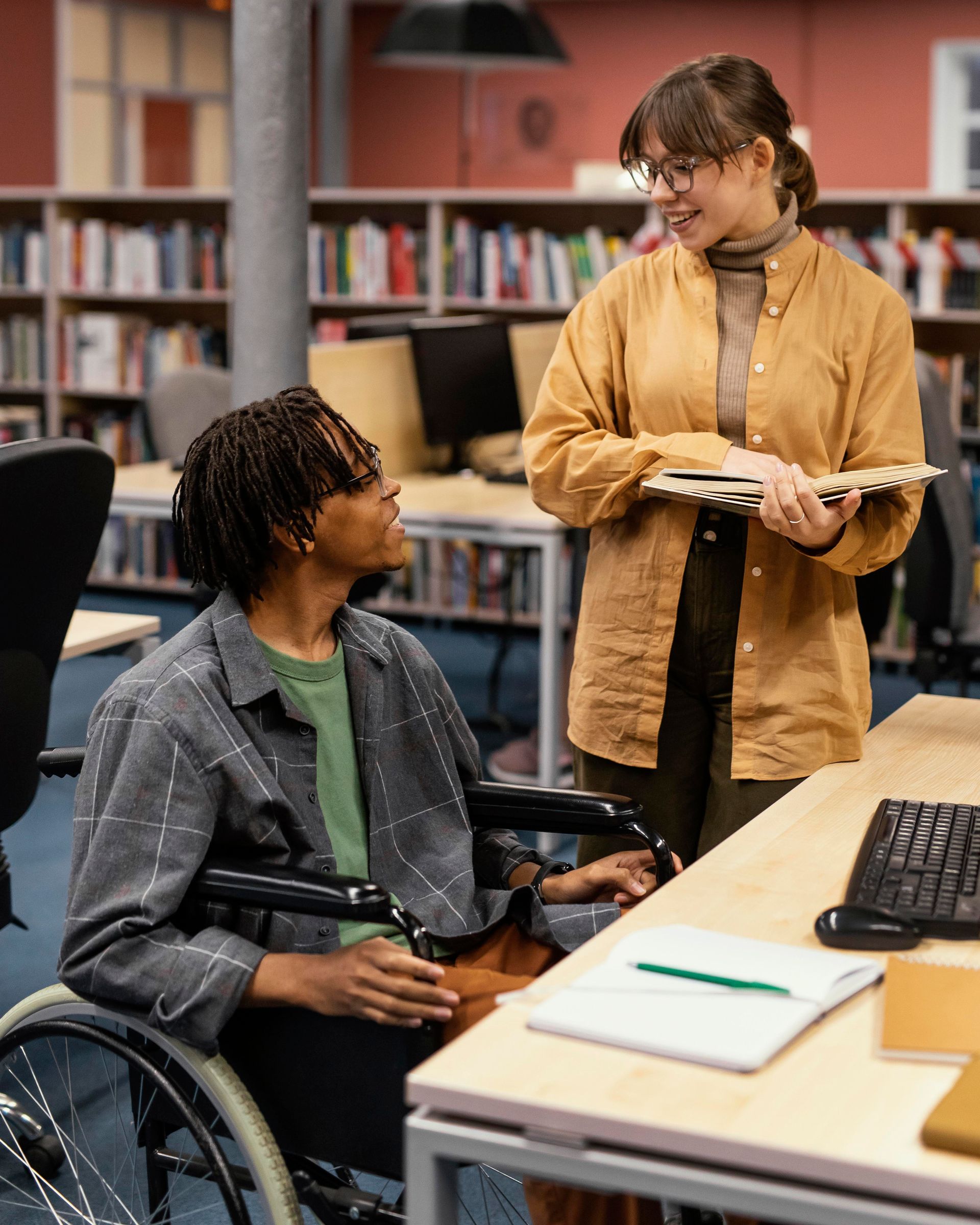 Two students in a library study area, one in a wheelchair, talking beside a desk with a keyboard and notebook.