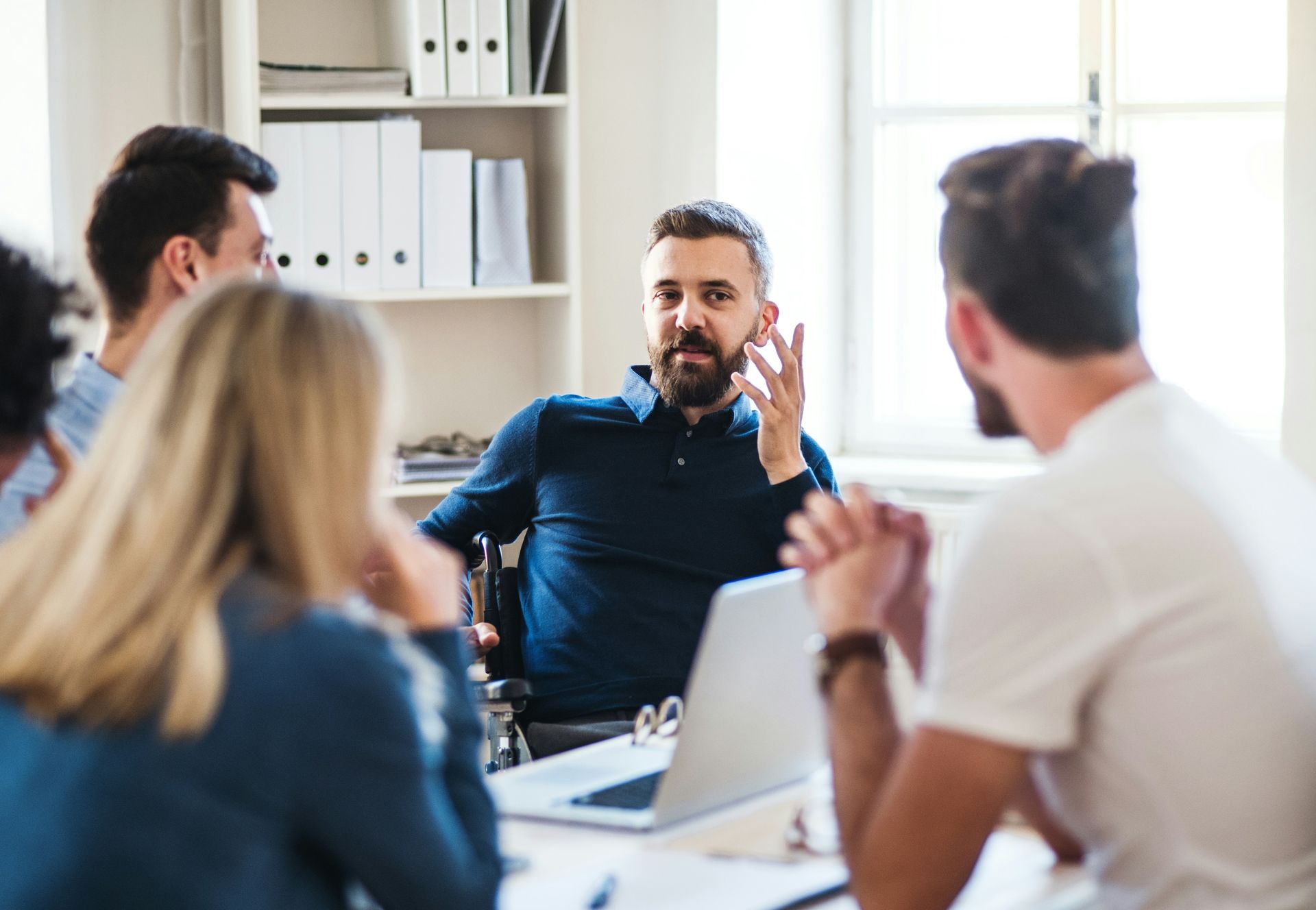 Small office meeting with a man speaking to colleagues around a table, laptops open.