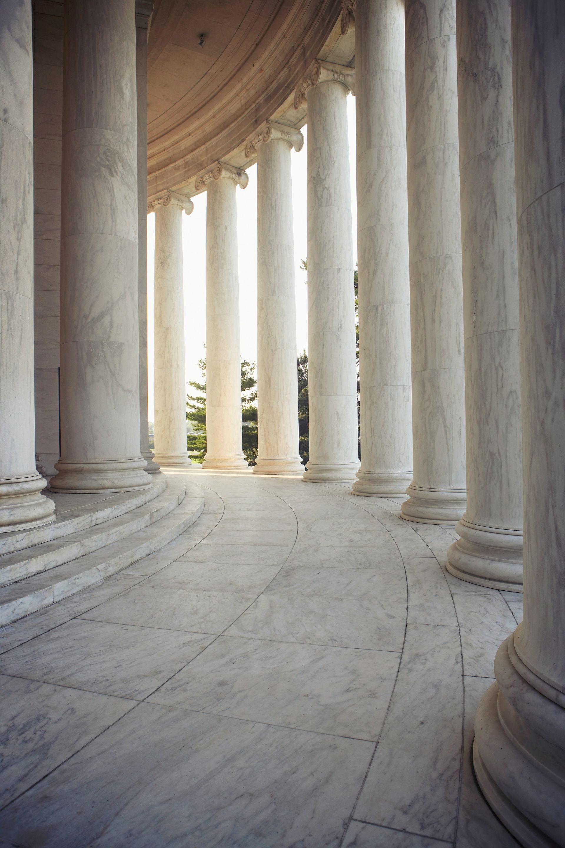 Curved colonnade of tall white marble columns with steps and sunlight filtering through openings