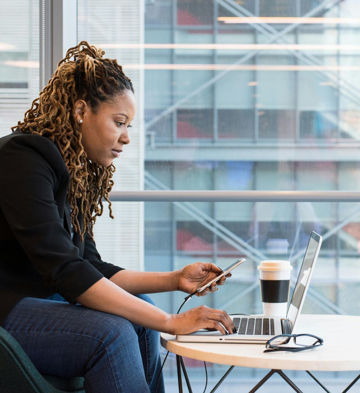 Woman working on a laptop and phone at a table by a large window, with coffee beside her