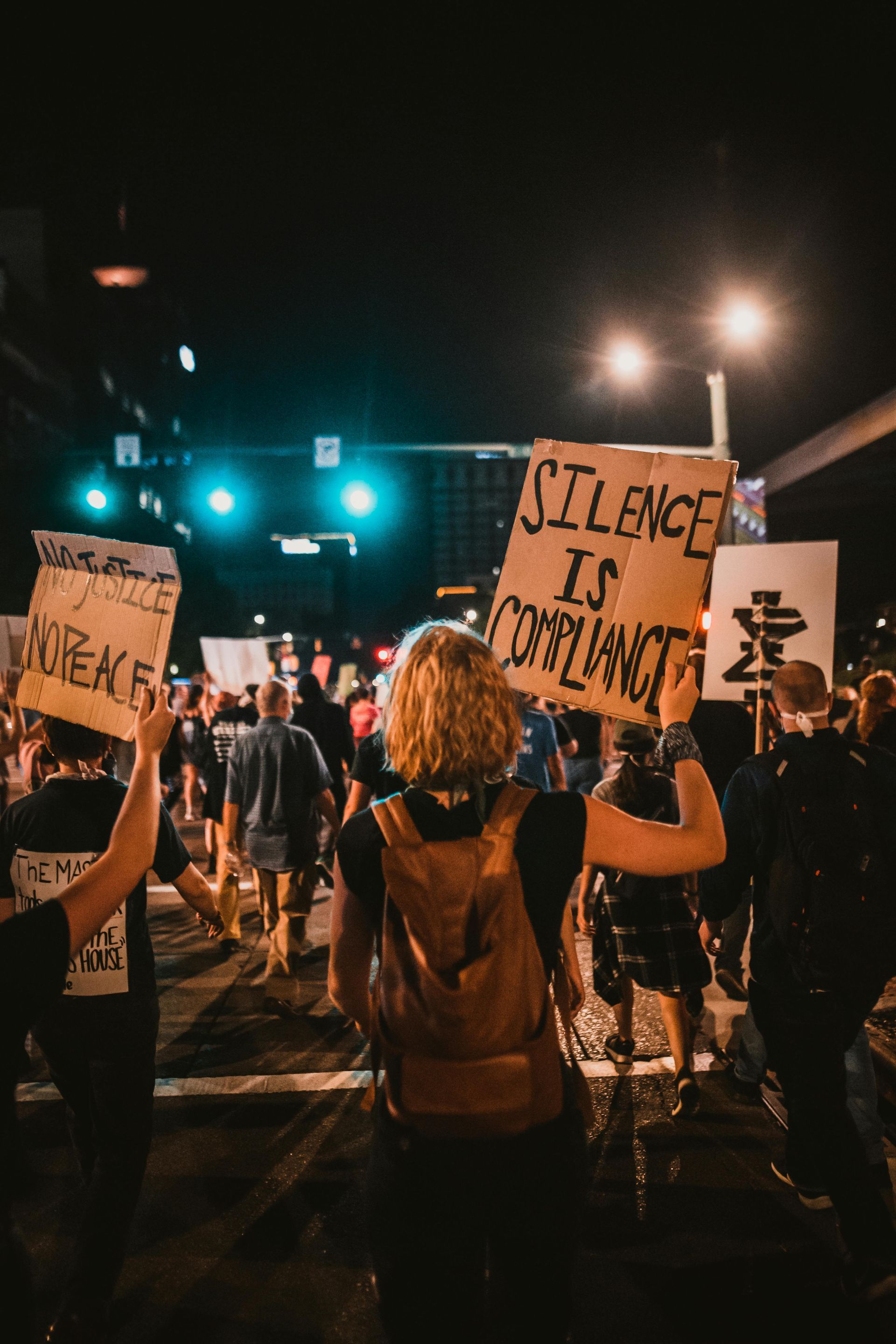 Protesters at night hold signs; one reads “Silence is complicity” in the center of the crowd.