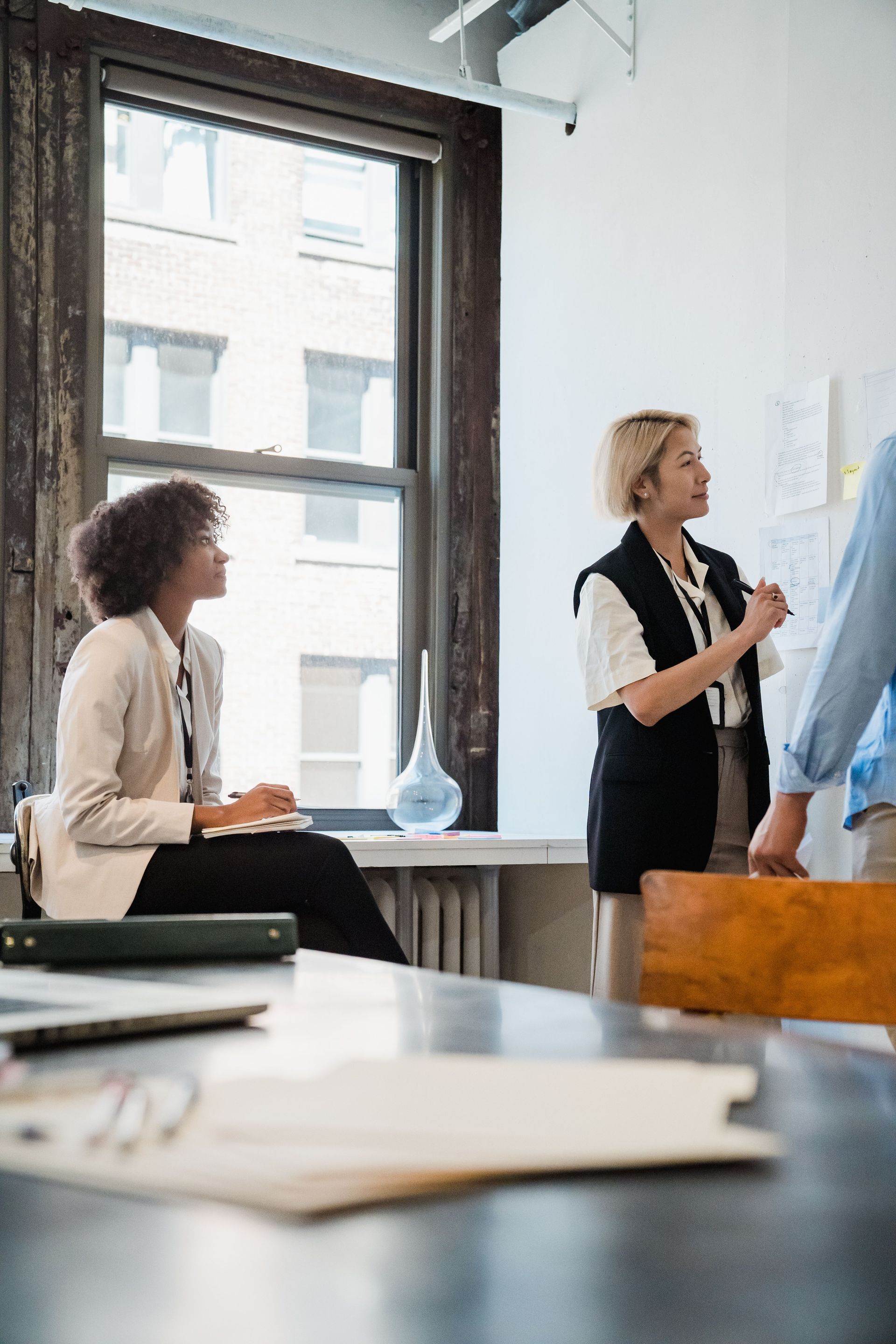 Two people in a bright office, one presenting a chart while the other listens near a window.