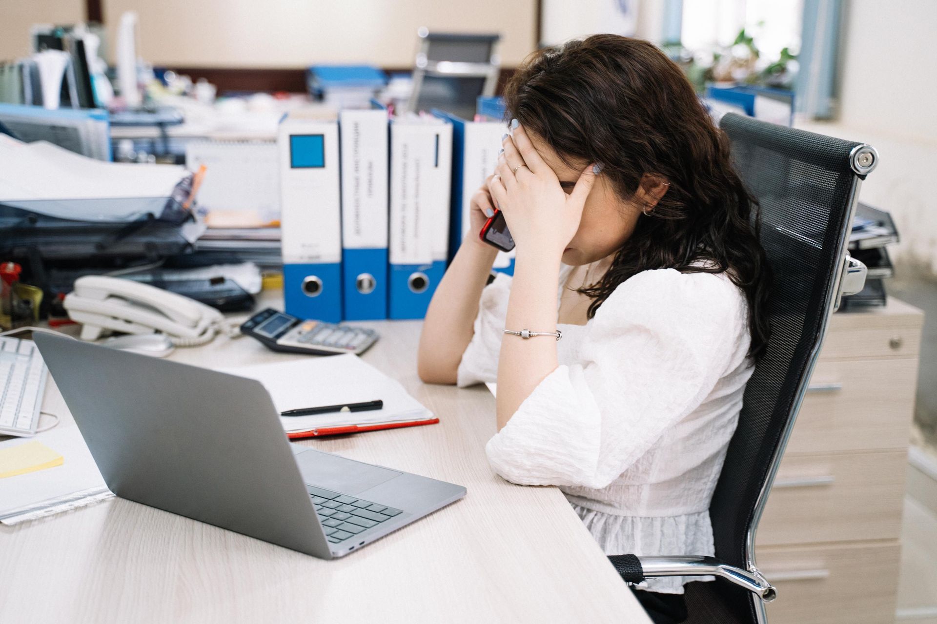 Frustrated office worker sitting at desk with laptop, hand covering face amid paperwork and binders