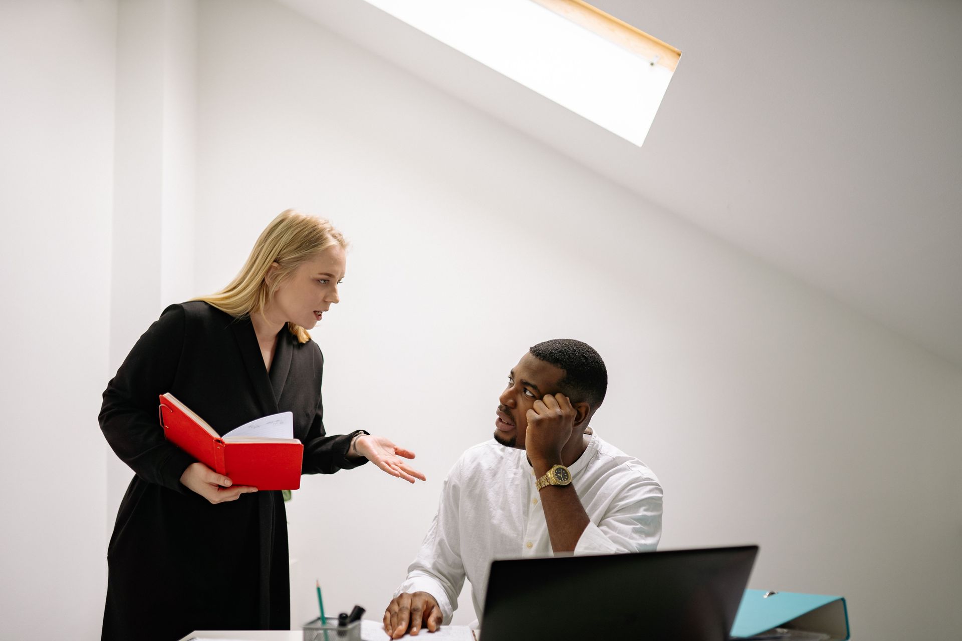 Two coworkers talking in a bright office, one holding red folders beside a laptop.