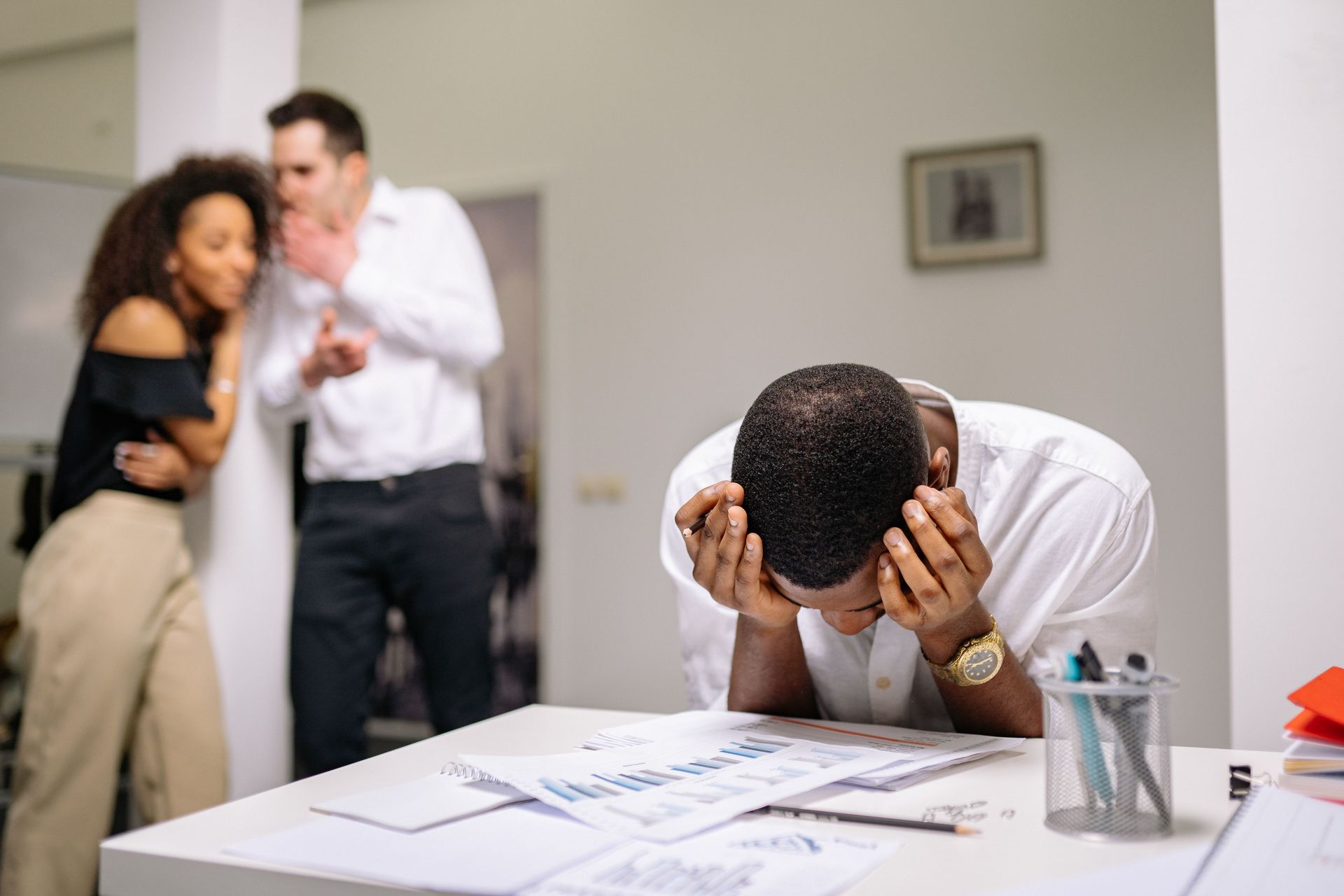 Stressed person at a desk covering their face while two blurred colleagues argue in the background.