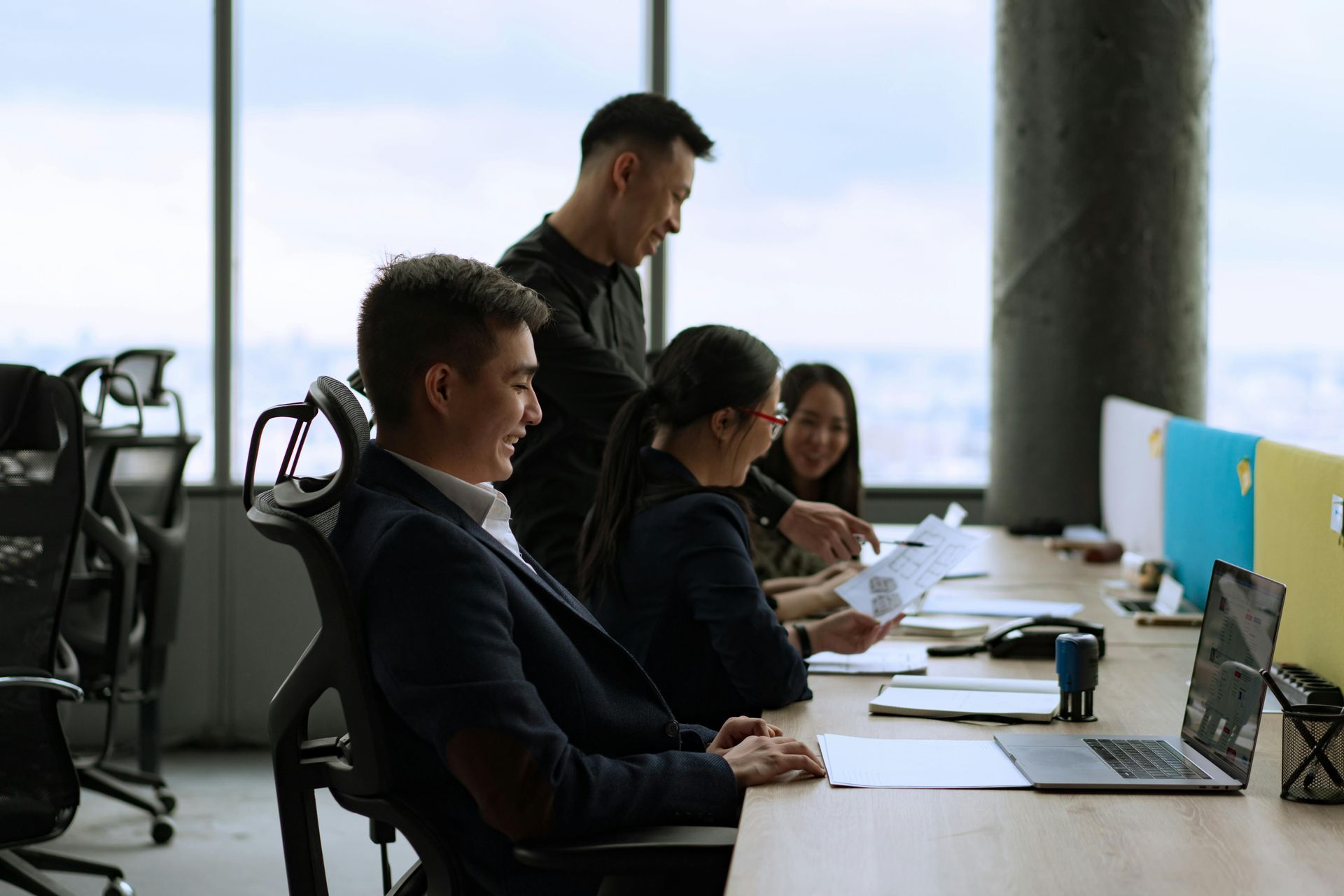 People working at a long desk in a bright office, focused on laptops and paperwork.