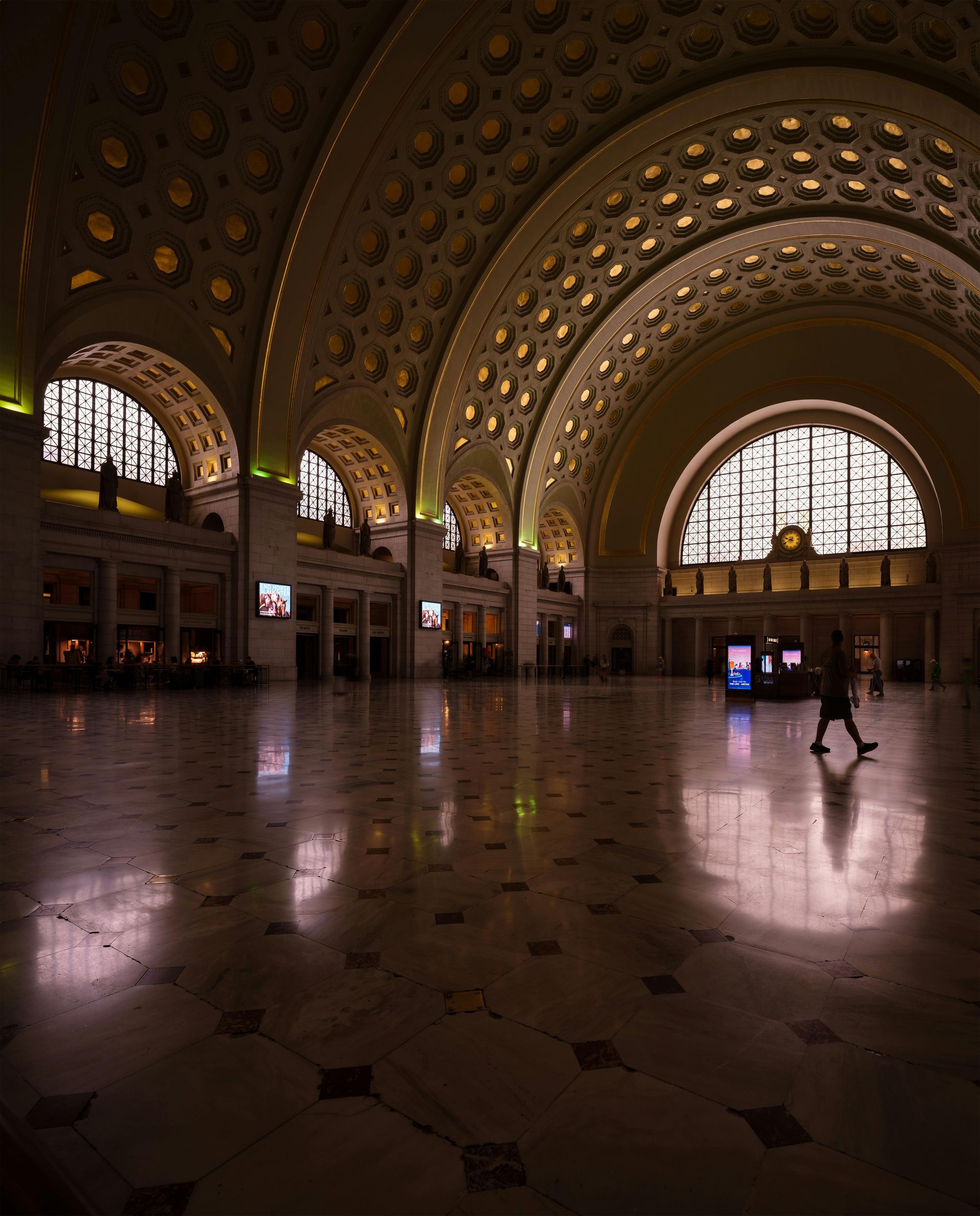 Grand, ornate train station hall with arched windows and reflective tiled floor, a few people walking inside