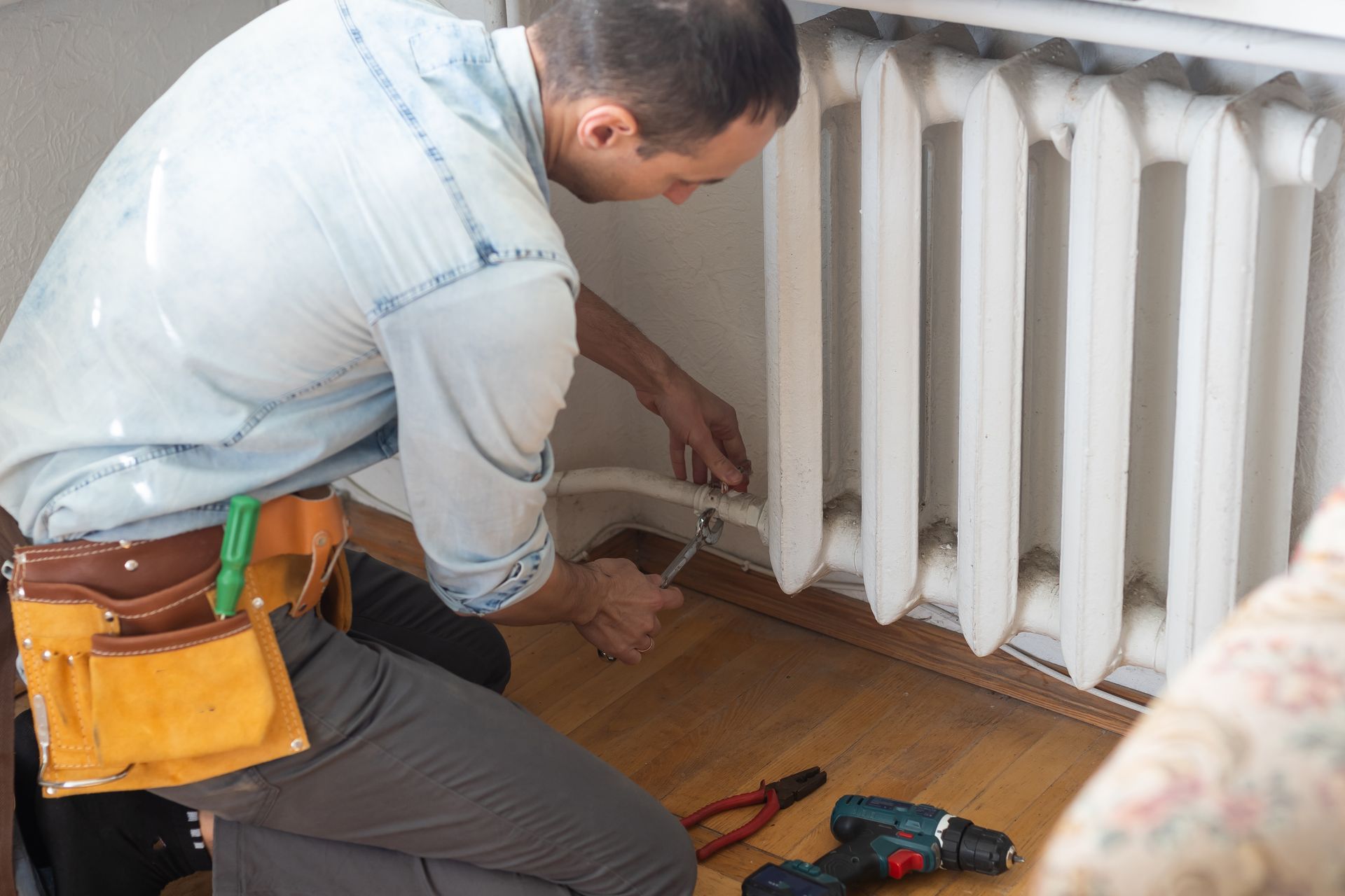 Technician repairing a white radiator near a wooden floor, with tools in a yellow pouch.
