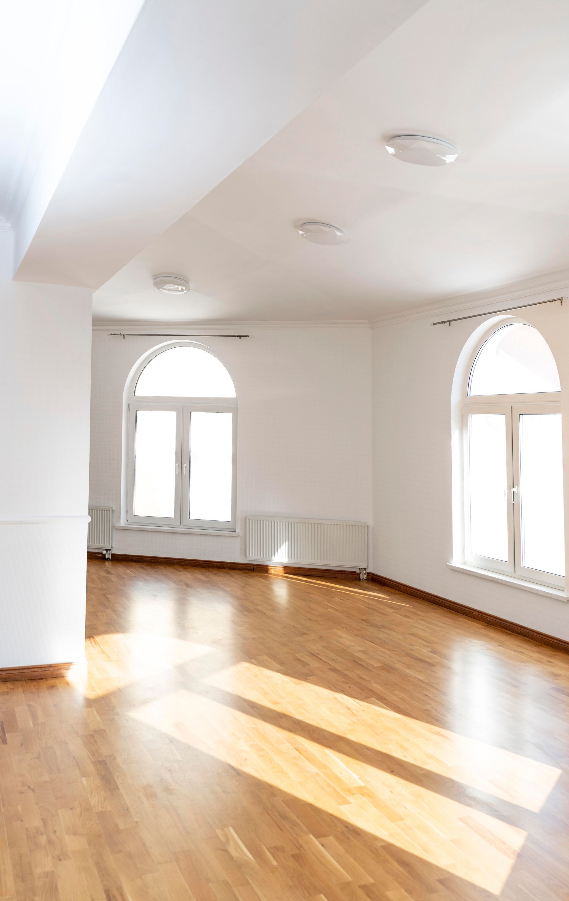 Bright empty room with hardwood floors, arched windows, and sunlight streaming across the floor.