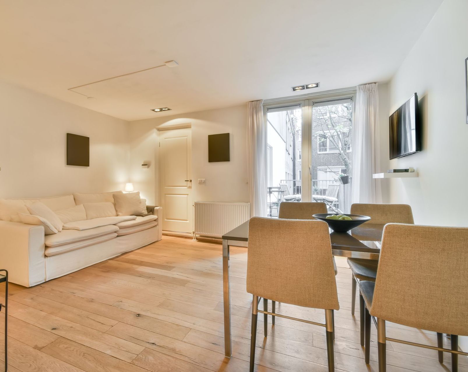 Bright living-dining room with white sofa, wooden floor, and a table by a window.