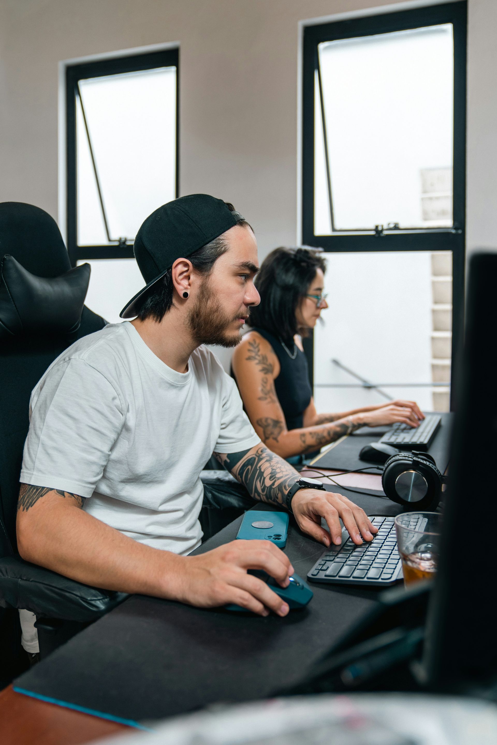 Two people working at computers in an office, with large windows and desks visible.