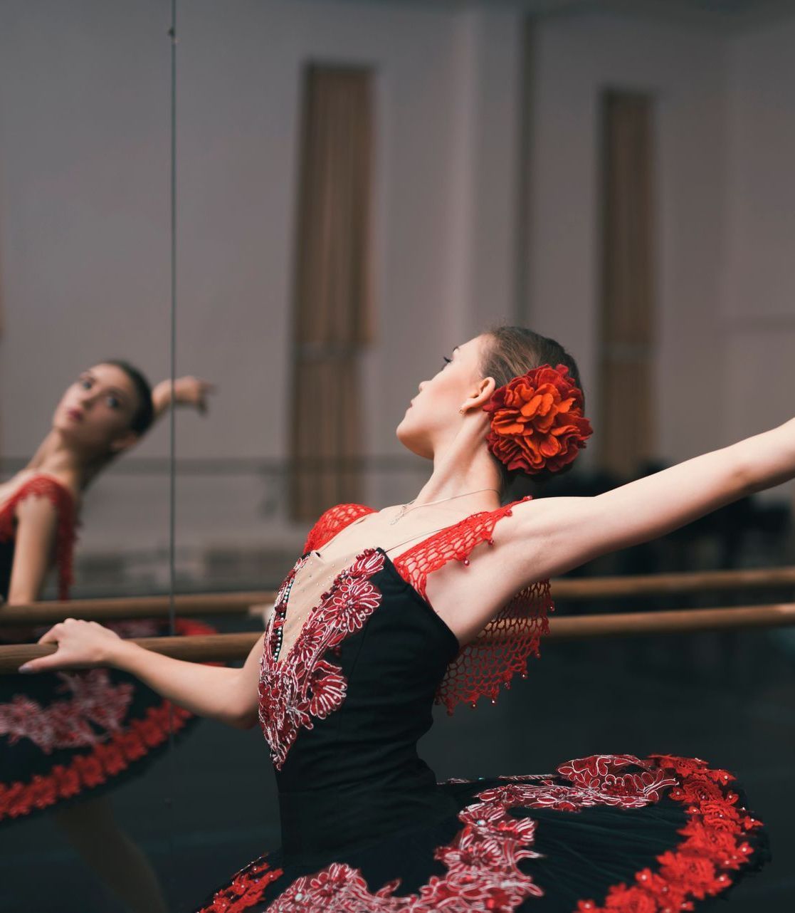 Ballet dancer in a red and black dress posing with arms outstretched in a studio, another dancer blurred behind