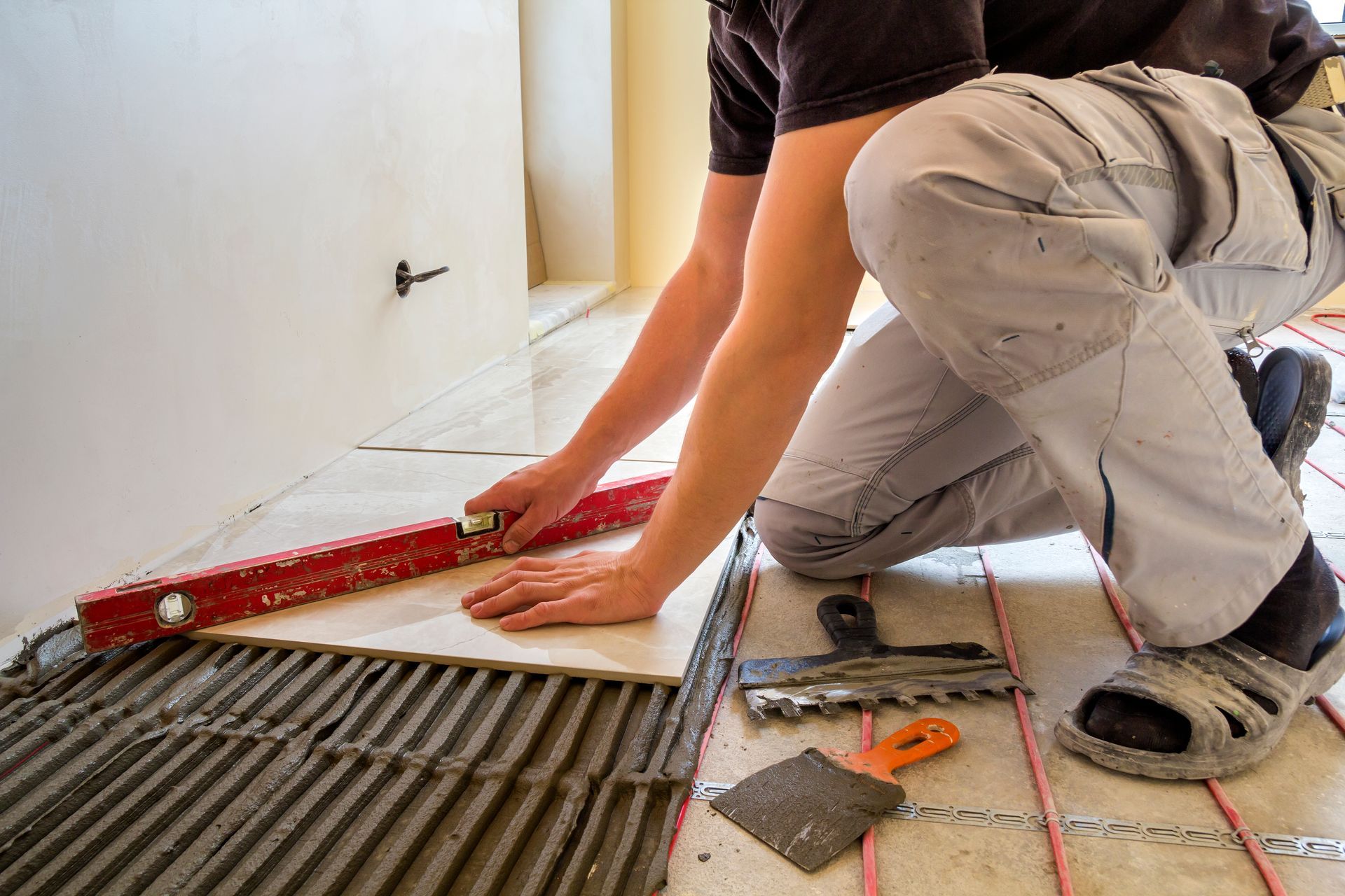 Worker installing tile floor with trowel, red level, and orange tools on a tiled surface