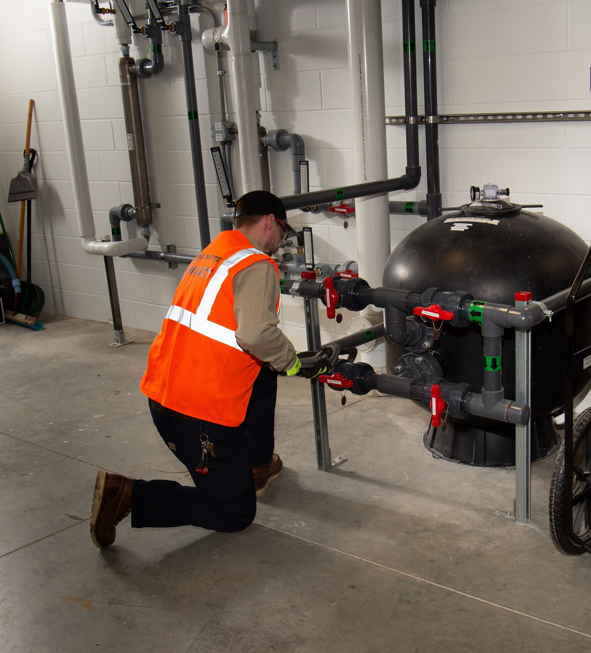Worker in orange safety vest kneels beside industrial piping and a large black tank in a utility room