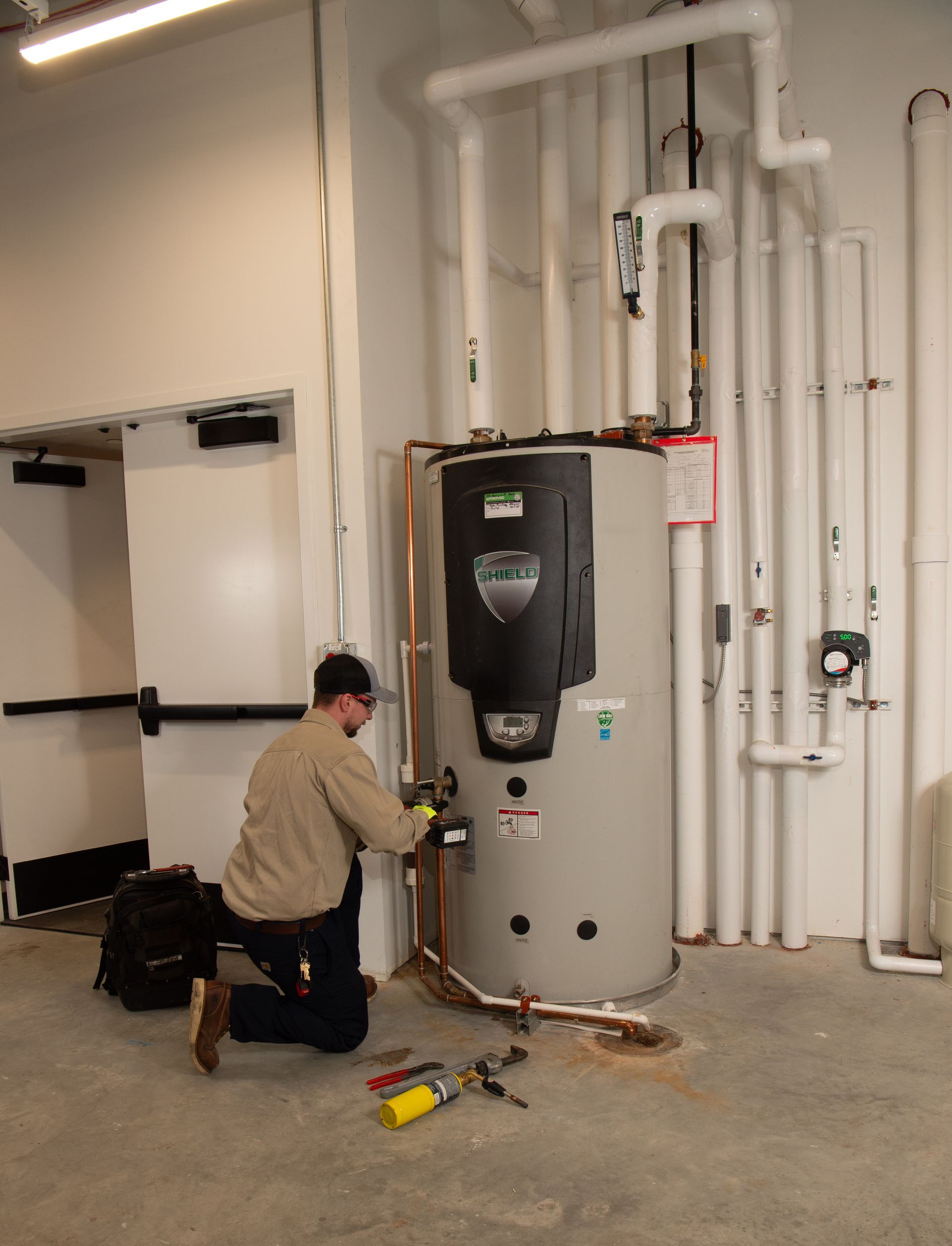 Technician kneels beside a large water heater in a utility room, working with tools and pipes.