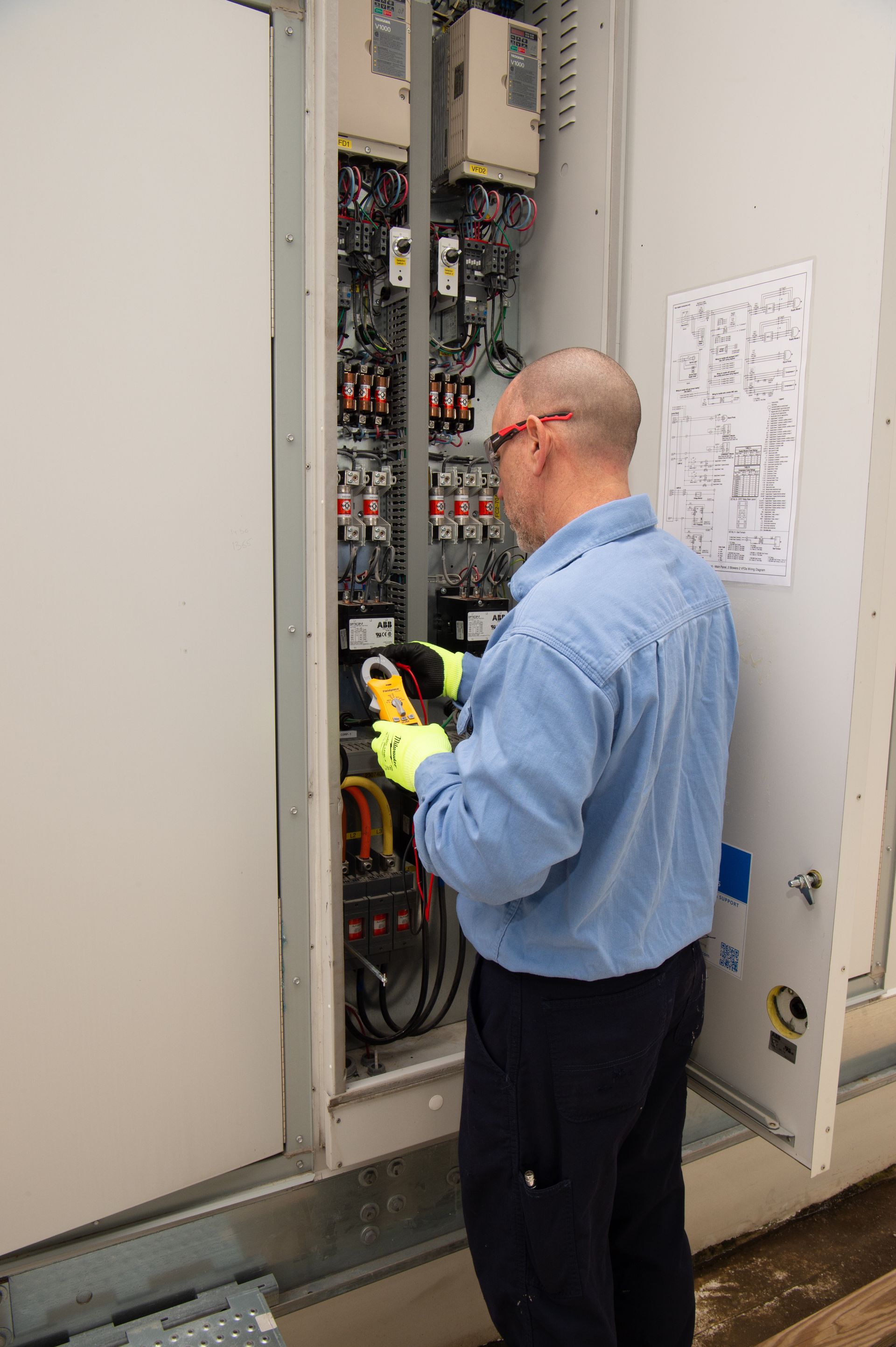 Technician in blue shirt inspecting wiring inside an open electrical cabinet, holding a yellow device.