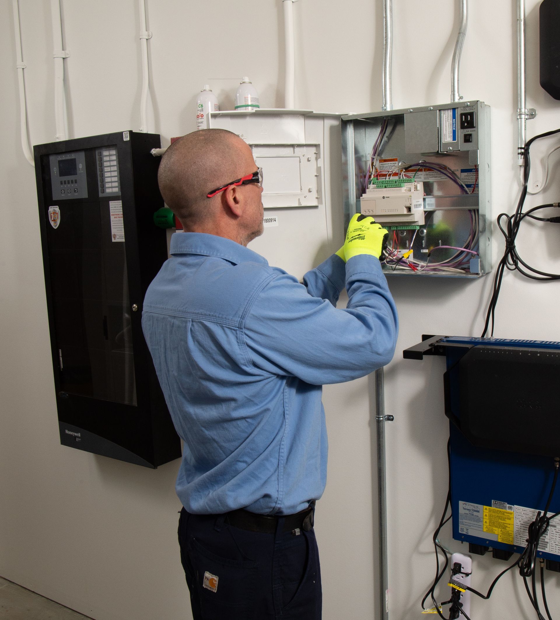 Technician in blue shirt and gloves inspecting an open electrical panel mounted on a wall