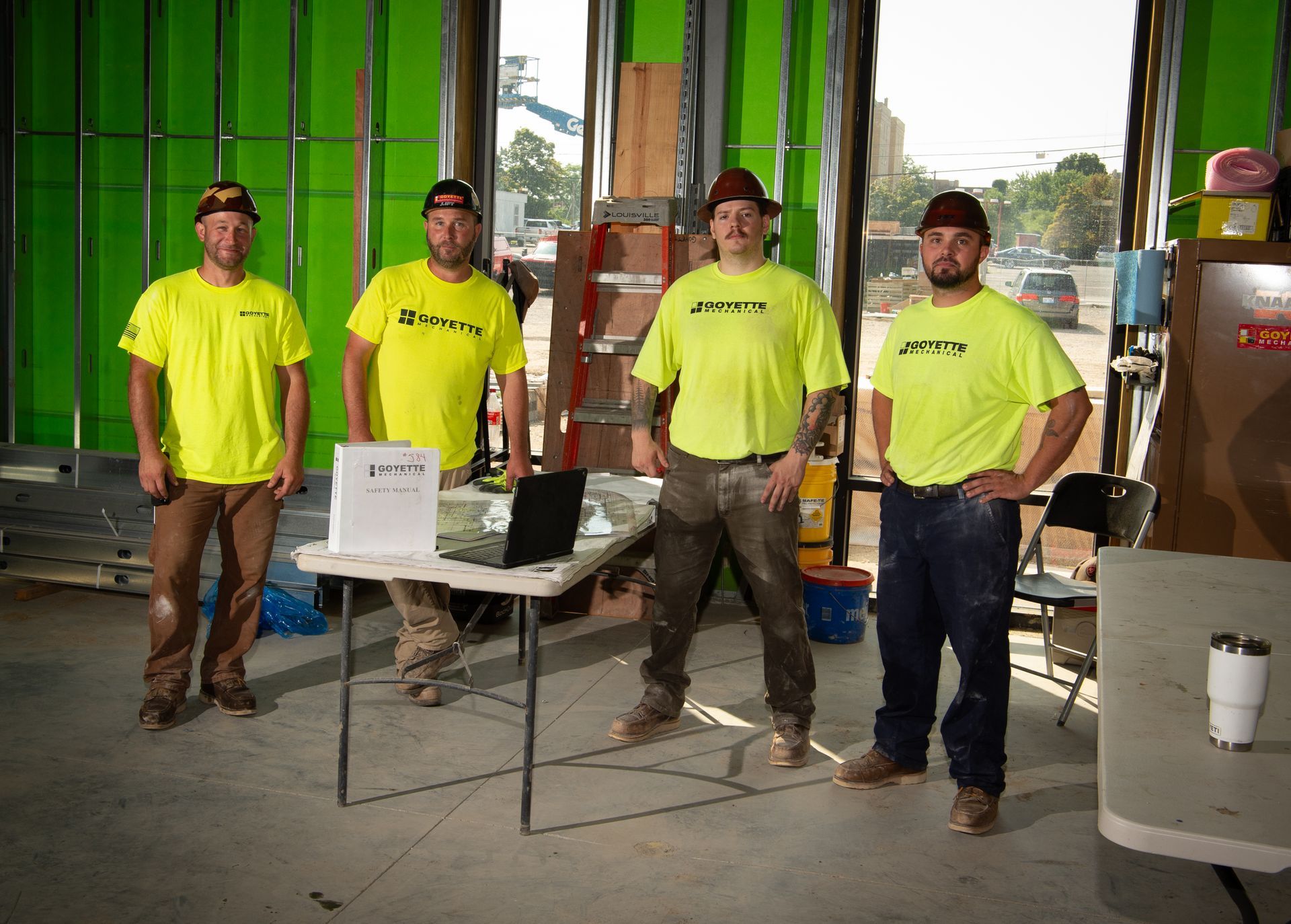 Four construction workers in yellow shirts and hats stand by a table in a building.