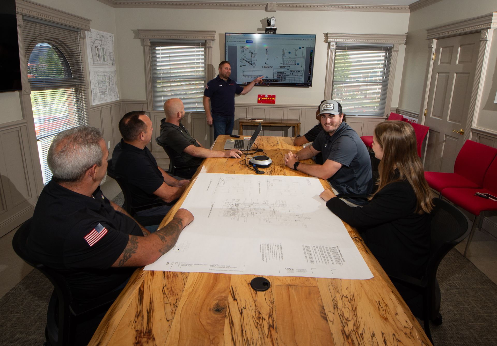 A group of people in a meeting room, reviewing blueprints and a presentation on a screen.