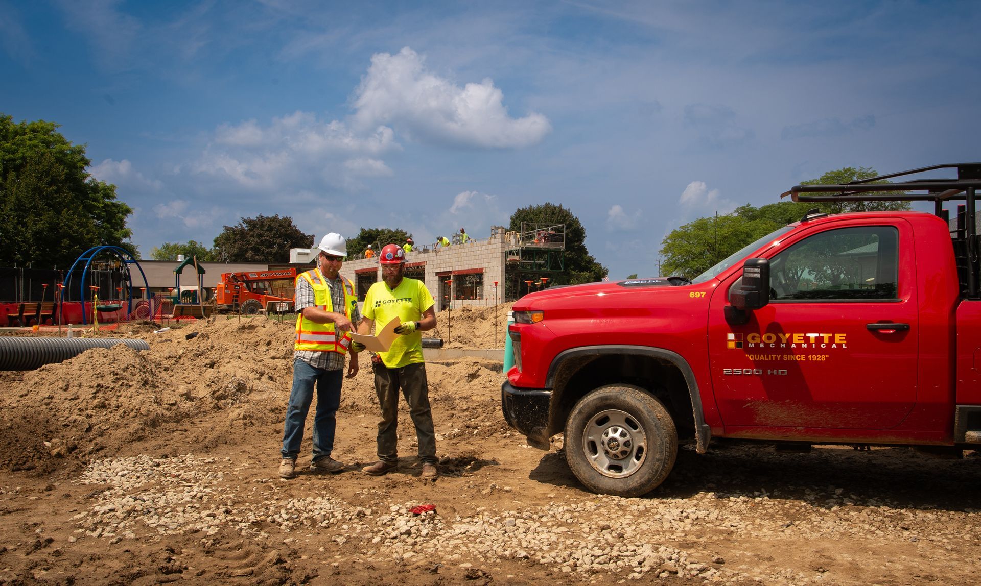 Two construction workers discuss plans beside a red pickup truck at a construction site on a sunny day.
