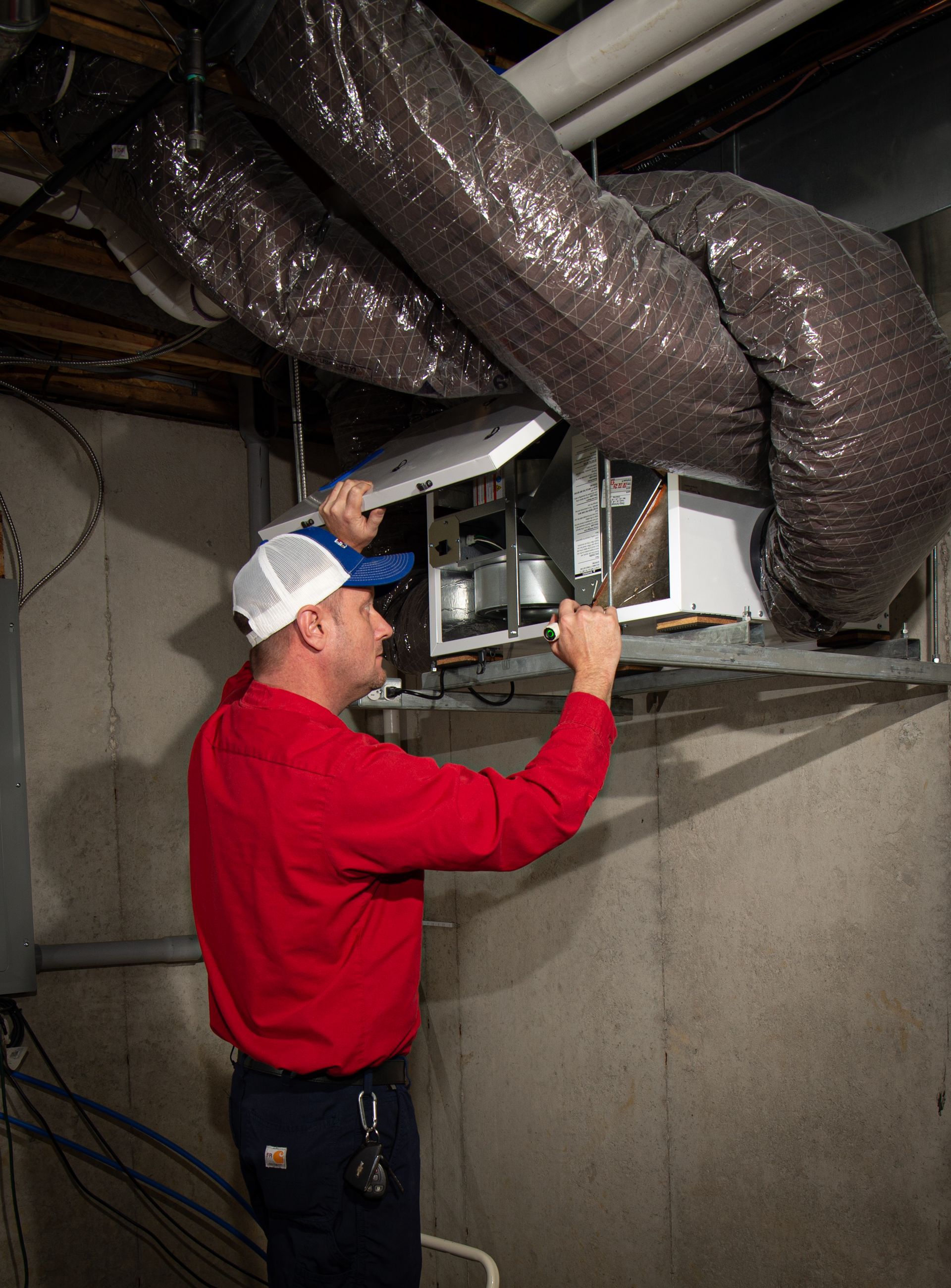 Technician in red shirt inspecting HVAC unit and ductwork in a basement ceiling area