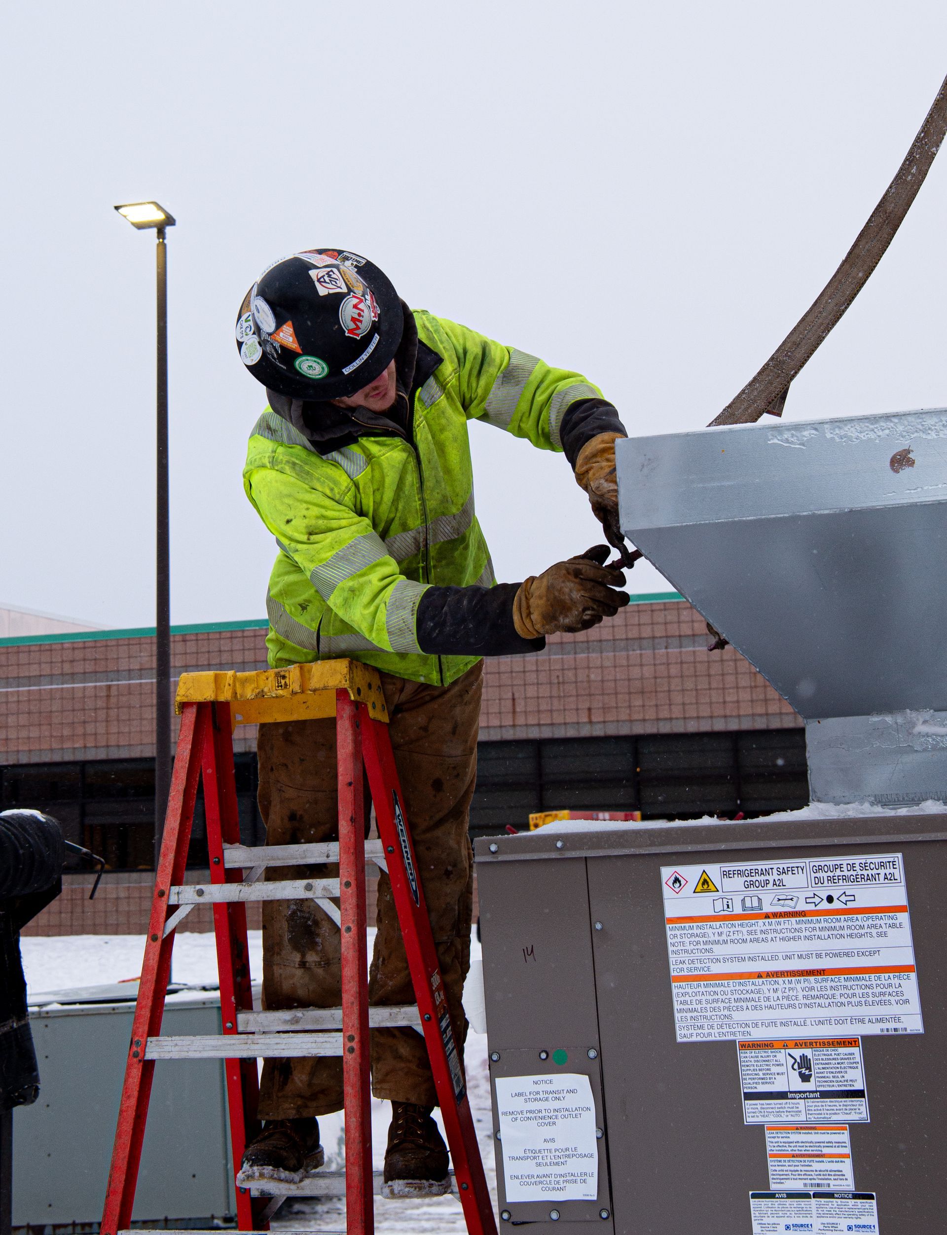 Person on a ladder installing ductwork on a snowy rooftop, wearing a reflective yellow jacket and work gloves.